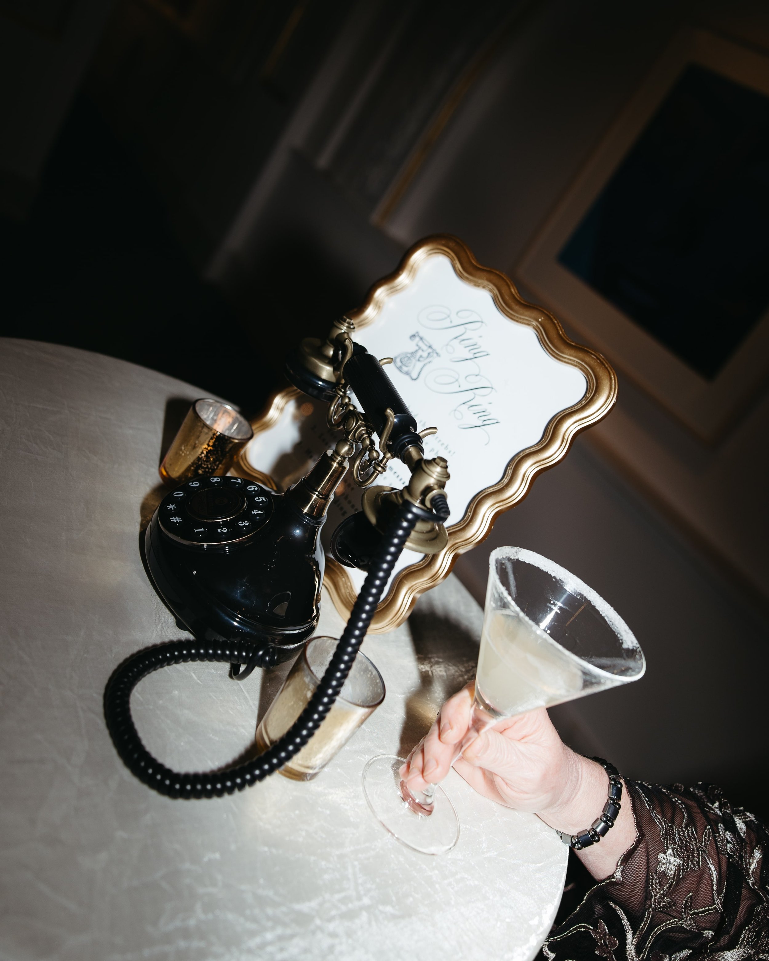 Creative wedding detail photo of a vintage black rotary phone guestbook display on a round white linen cocktail table. A framed sign reading “Ring Ring Leave a Message” sits behind the phone, alongside votive candles and a guest’s hand holding a suga