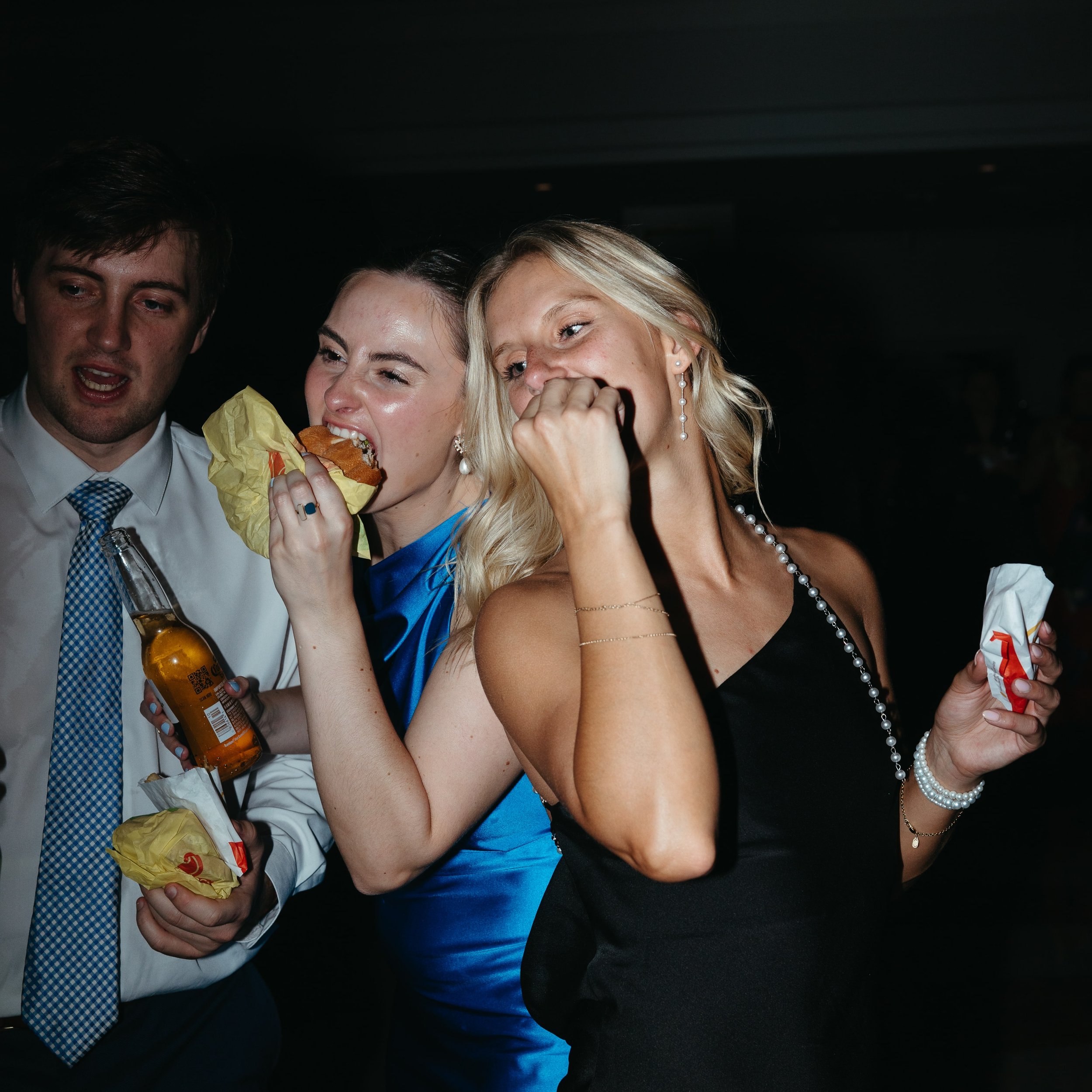 Documentary wedding reception photo by Kathleen Baker Photography showing guests indulging in late-night fast food. Two women in formal gowns—one in bright blue satin, the other in black with pearls—laugh while eating McDonald’s cheeseburgers and fri