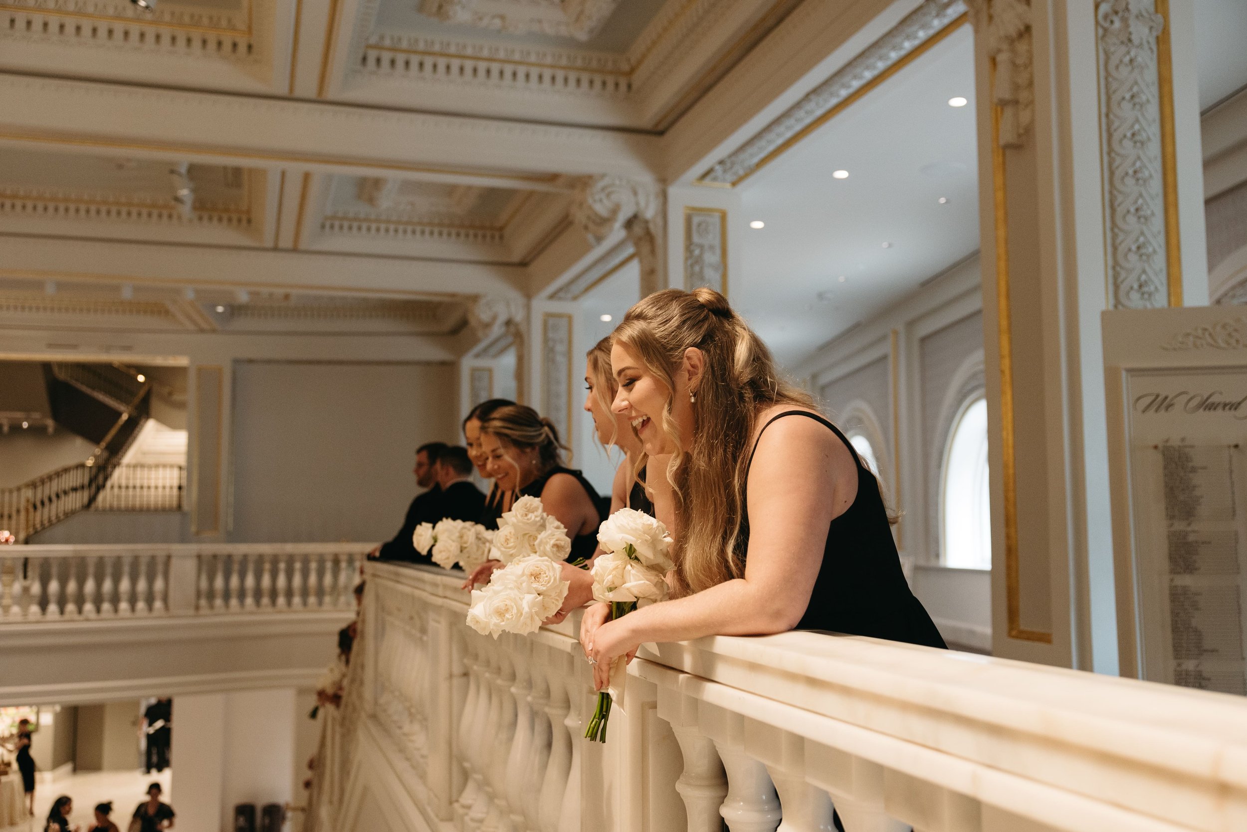 Documentary wedding photography of bridesmaids in black dresses holding ivory rose bouquets, leaning over an ornate white marble balcony railing in a grand ballroom reception venue with classical architectural details, gold trim, and arched windows i
