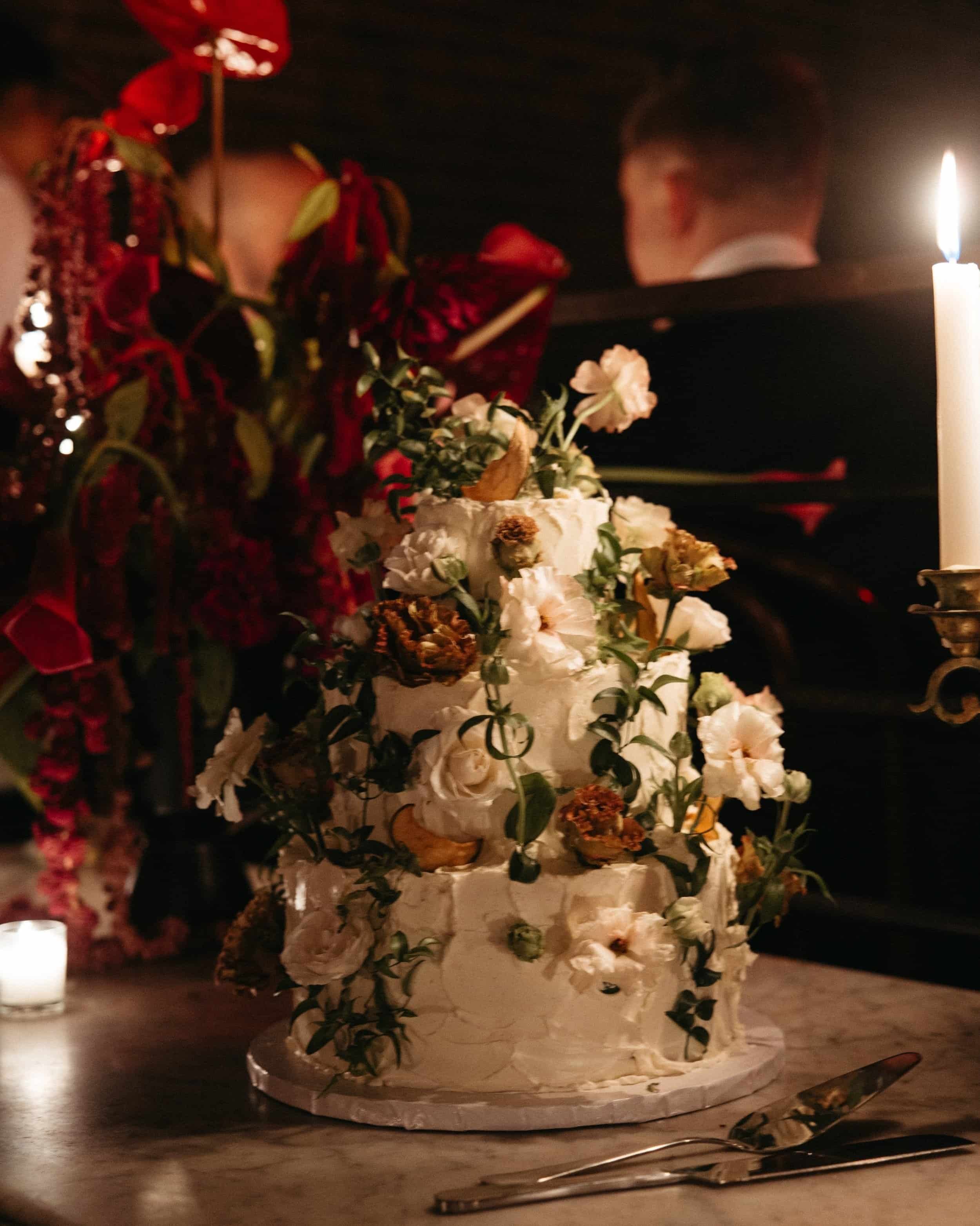 A three-tiered wedding cake sits on a marble table, illuminated by the warm glow of candlelight at Sadelle’s in New York City. The cake has an artfully rustic buttercream finish, with delicate swirls and textured edges. Fresh flowers in soft ivory, b