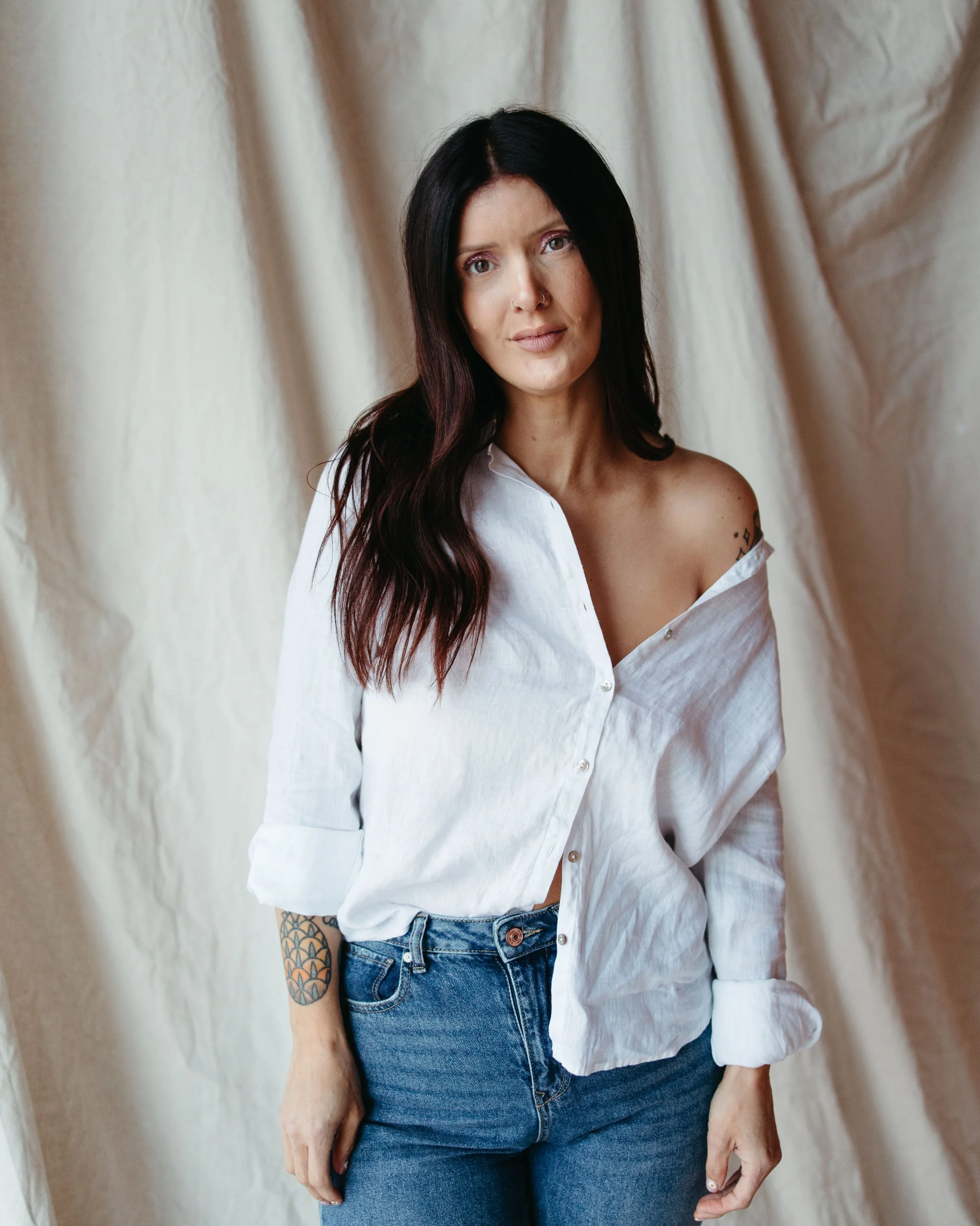 A softly lit headshot of the esthetician looking directly at the camera with a gentle smile. She stands in front of a neutral-toned fabric background, her long dark hair parted in the middle. Her linen shirt is slightly off one shoulder, adding a cas