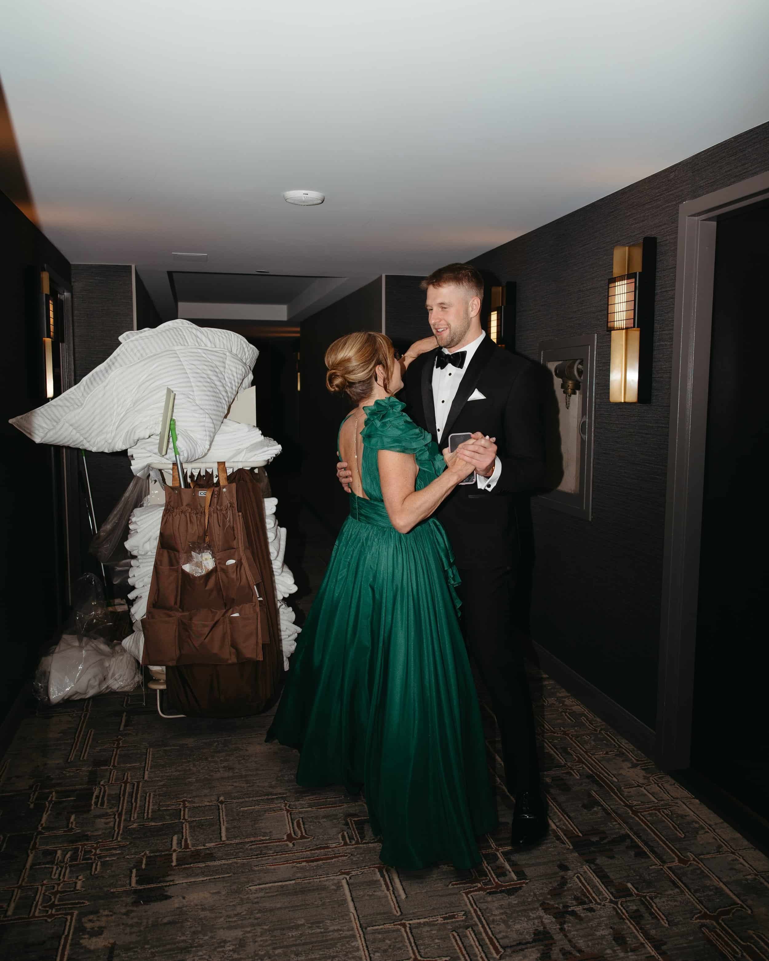 In the hallway of the Grand Hyatt Washington, the groom shares a sweet, impromptu dance practice with his mother before the ceremony. He wears a sharp black tuxedo while she stuns in a flowing emerald green gown. Behind them, a stack of hotel linens
