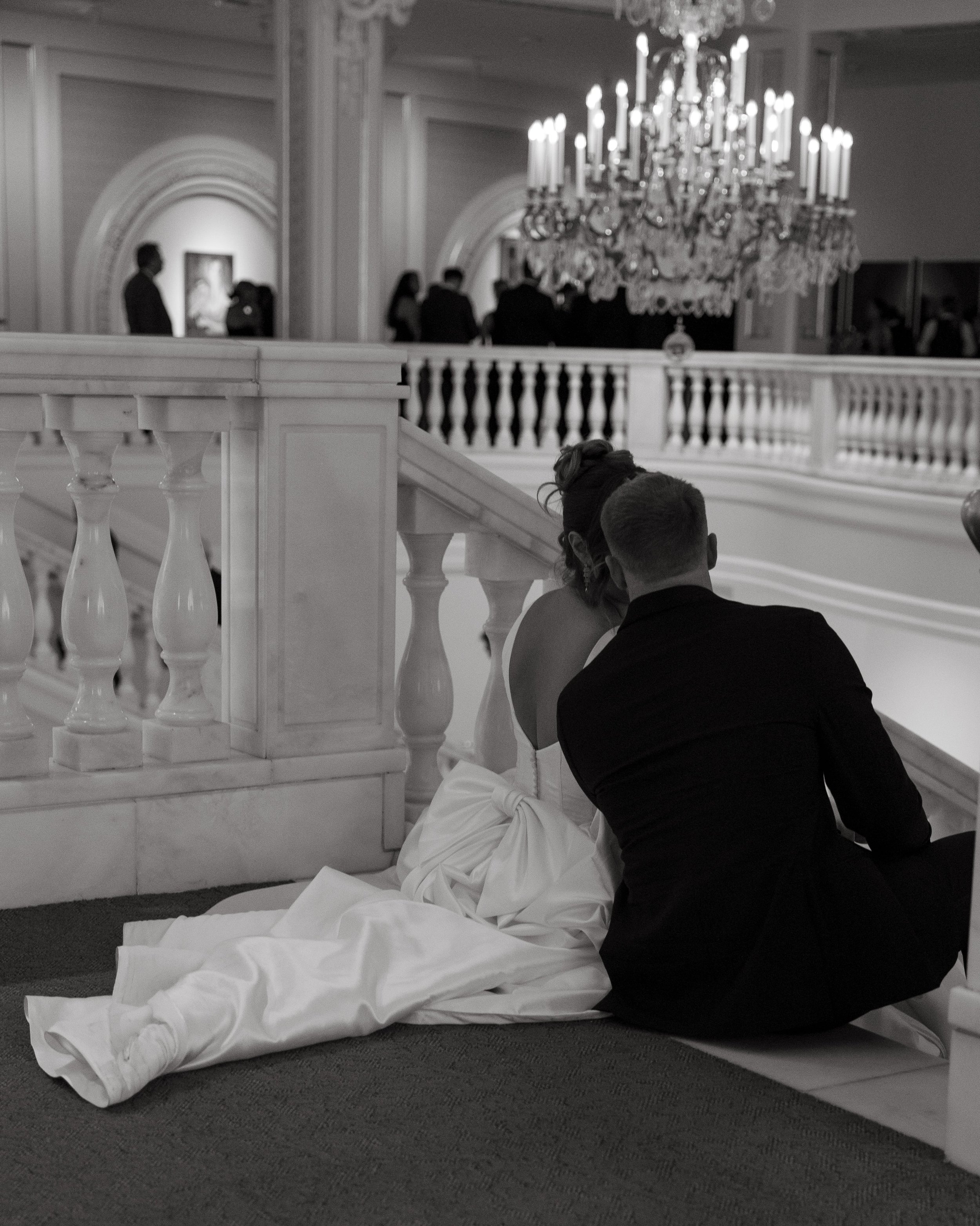 Black and white documentary wedding photograph of bride and groom sitting side by side on white marble stairs in an elegant reception venue with grand chandeliers and classical architecture. The bride wears a satin gown with a large bow detail on the