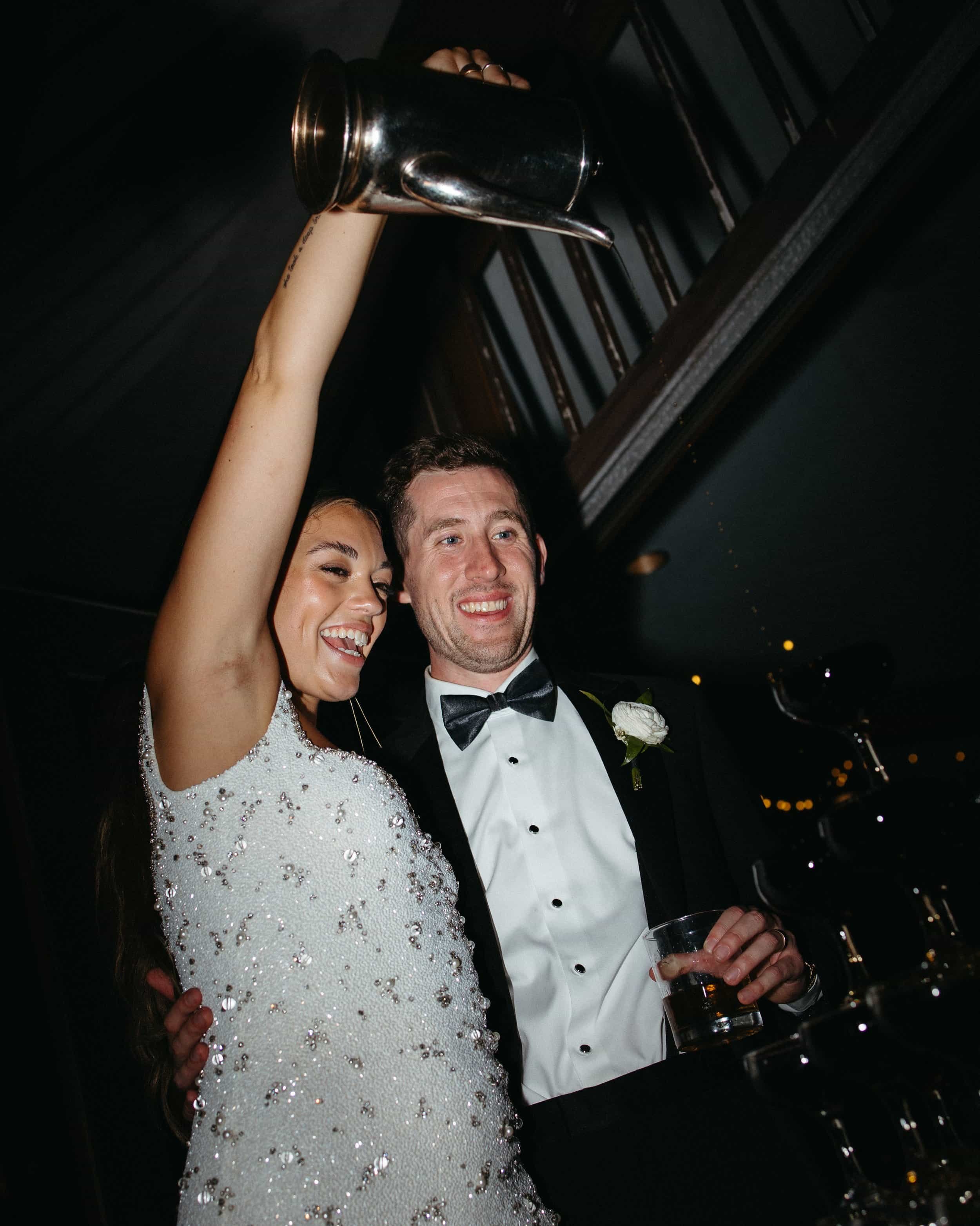 The bride and groom laugh as they pour espresso martini on an espresso martini tower at Winterthur Museum, celebrating in a high-end and glamorous reception setting. Bride is wearing a custom beaded reception mini dress and groom is wearing a traditi