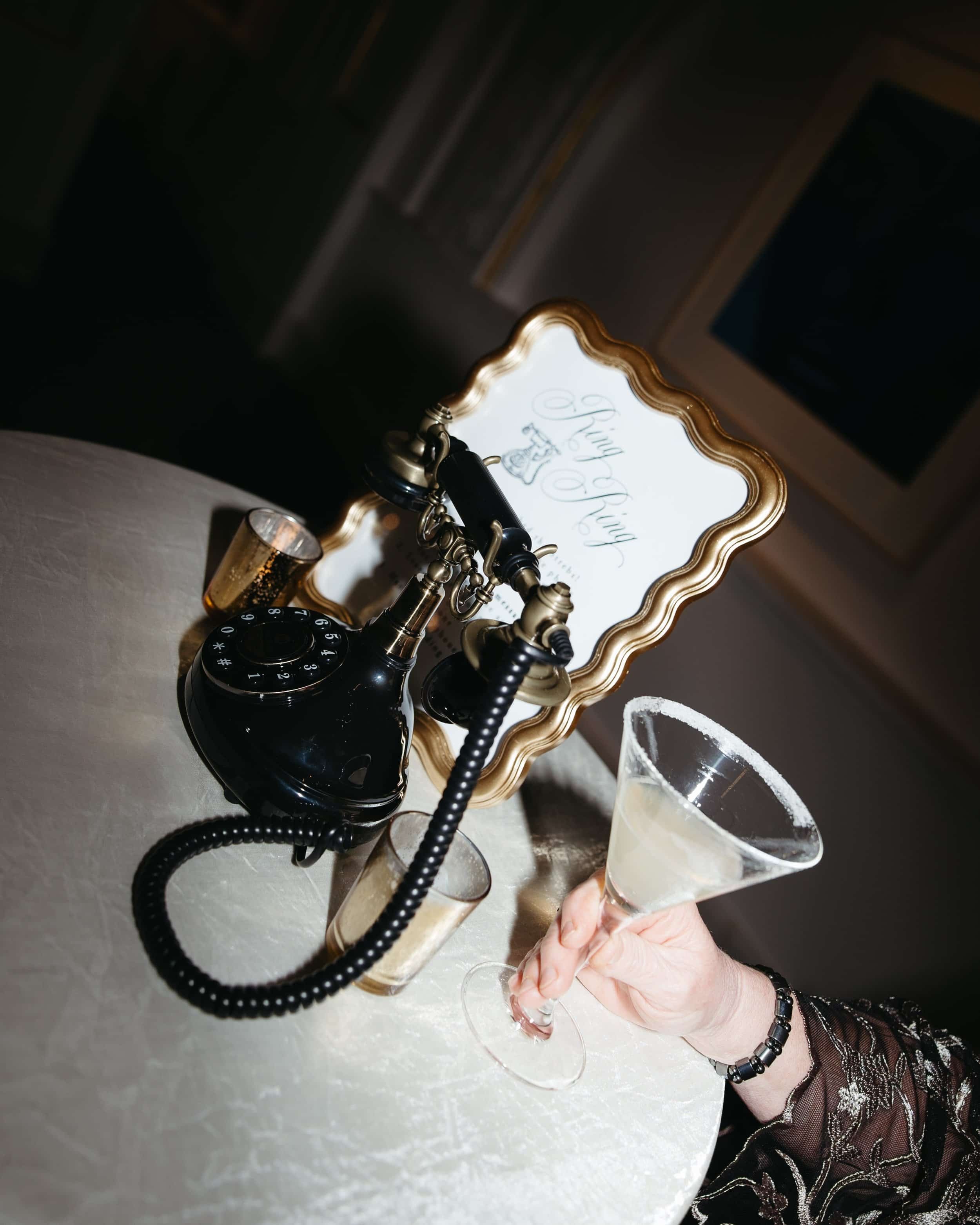 A guest’s hand holds a martini glass beside a vintage-style guestbook phone labeled “Ring Ring.” A moody, flash-lit image captured during cocktail hour at the National Museum of Women in the Arts. Kathleen is a luxury wedding photographer offering do