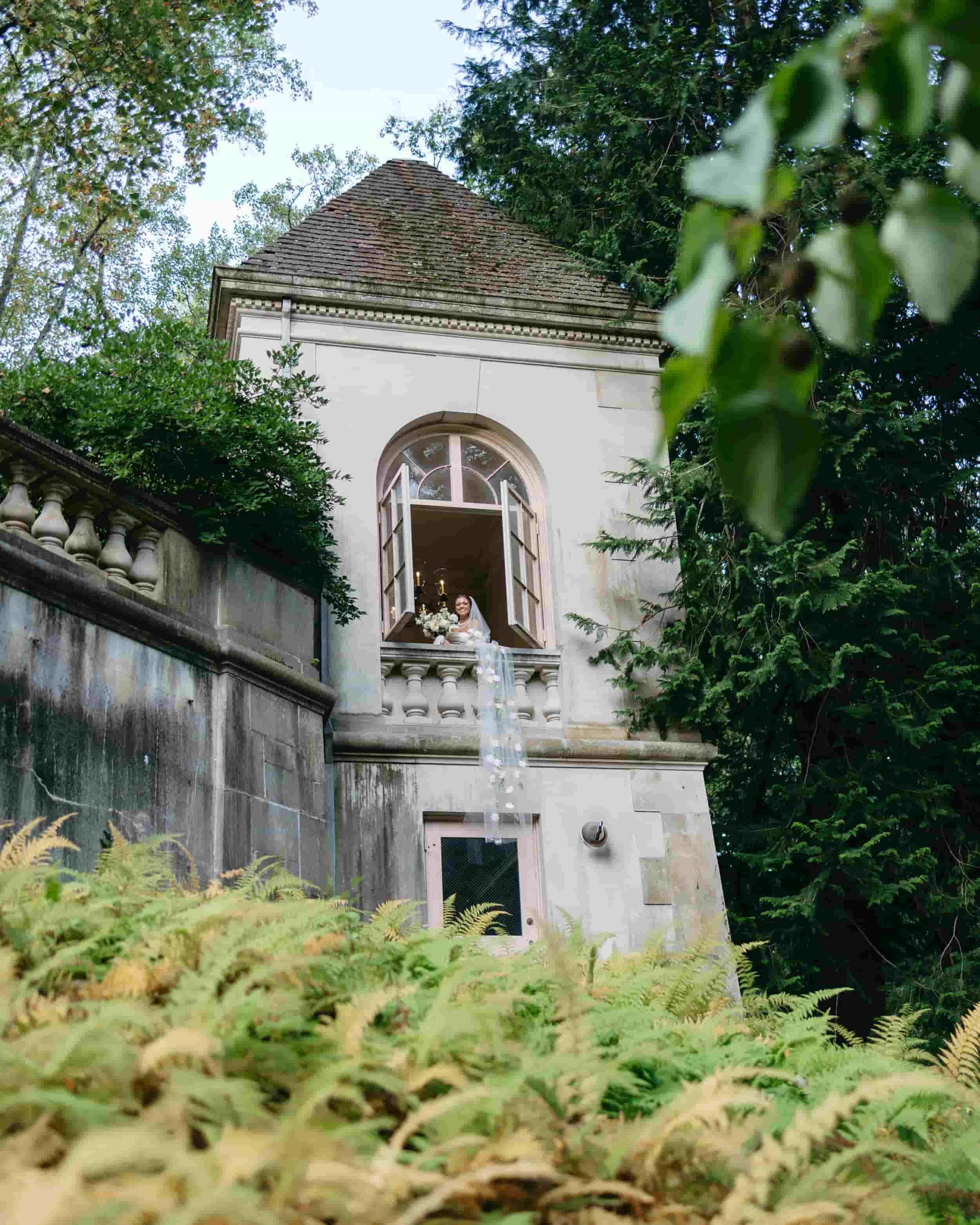 A romantic shot of the bride leaning out of a second-story window at Winterthur Museum, her veil cascading dramatically over the edge of the building. The surrounding greenery frames the moment beautifully. Kathleen is a luxury wedding photographer o