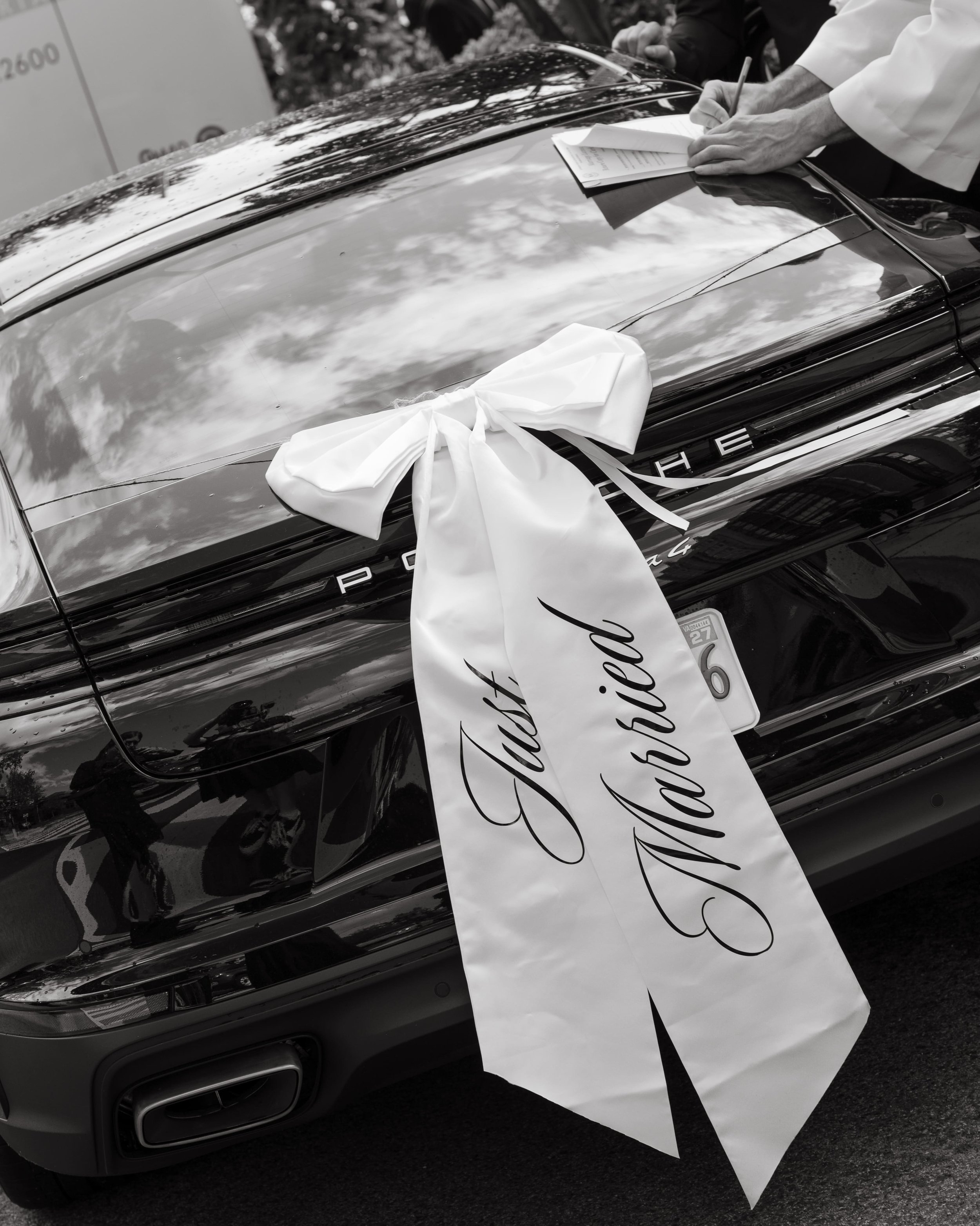 Black and white detail photo of a black Porsche decorated with a large white satin bow and long “Just Married” ribbon on the trunk. A man’s hand holds a pen signing paperwork on the back of the car. Captured outside a Washington DC wedding venue. Edi