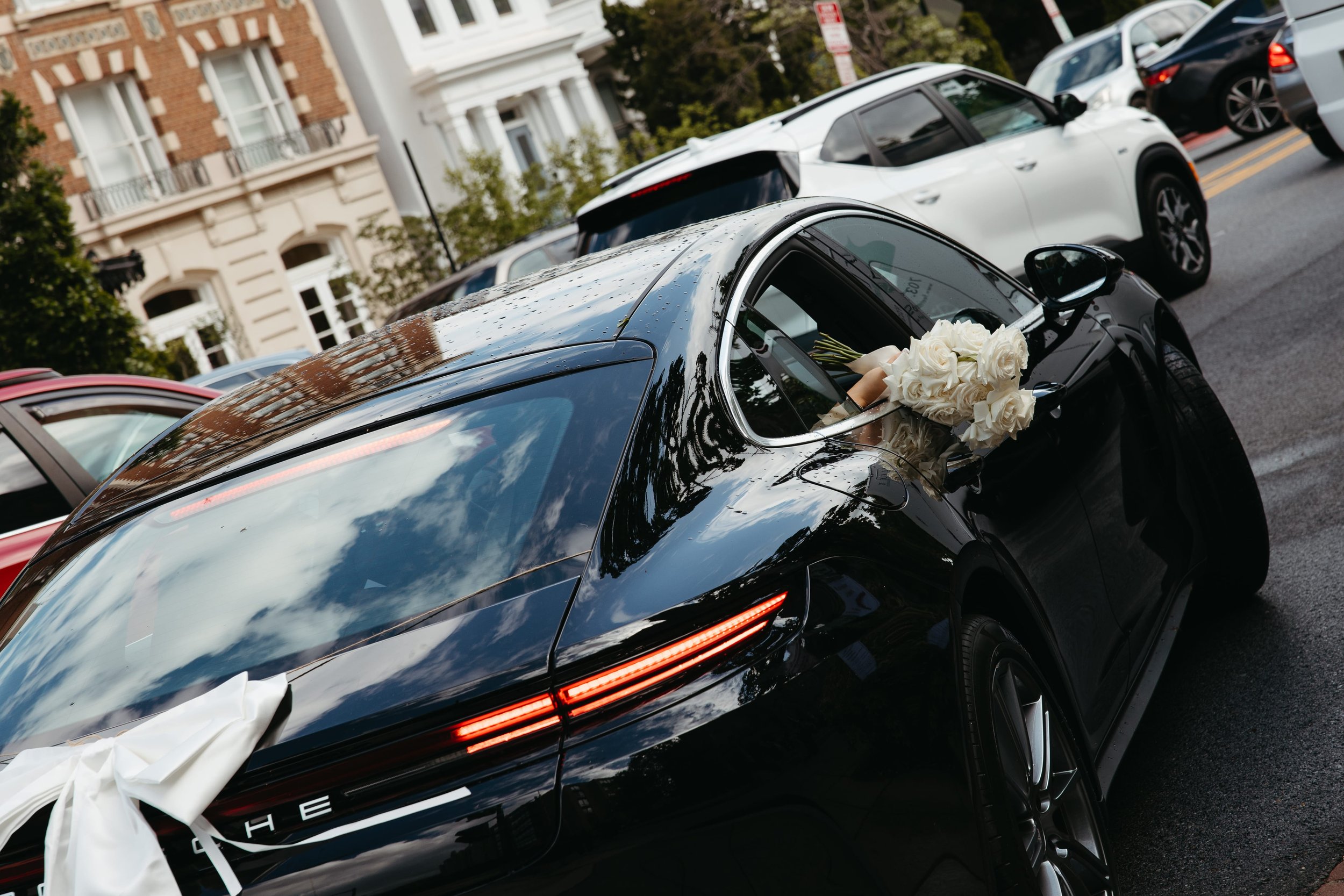 Editorial wedding photography of a black Porsche with white roses tucked into the side mirror and a large white satin bow on the trunk. The car drives away through a city street lined with parked cars and historic architecture in Washington DC. Docum