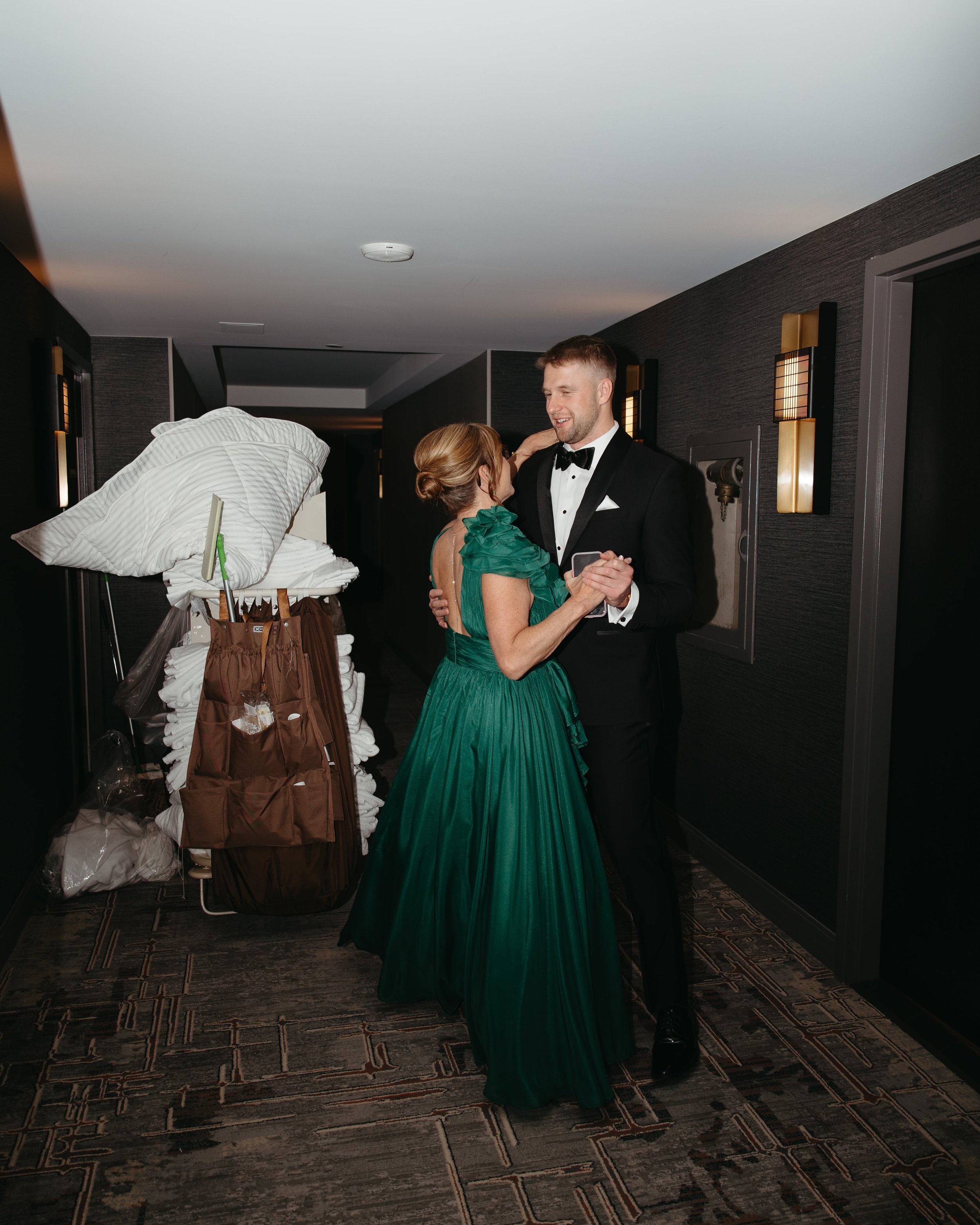 Documentary wedding photo of a groom in a classic black tuxedo with a white pocket square and bow tie, dancing with his mother wearing a vibrant emerald green gown with ruffle details. Captured in a dimly lit hotel hallway in Washington DC, with brow