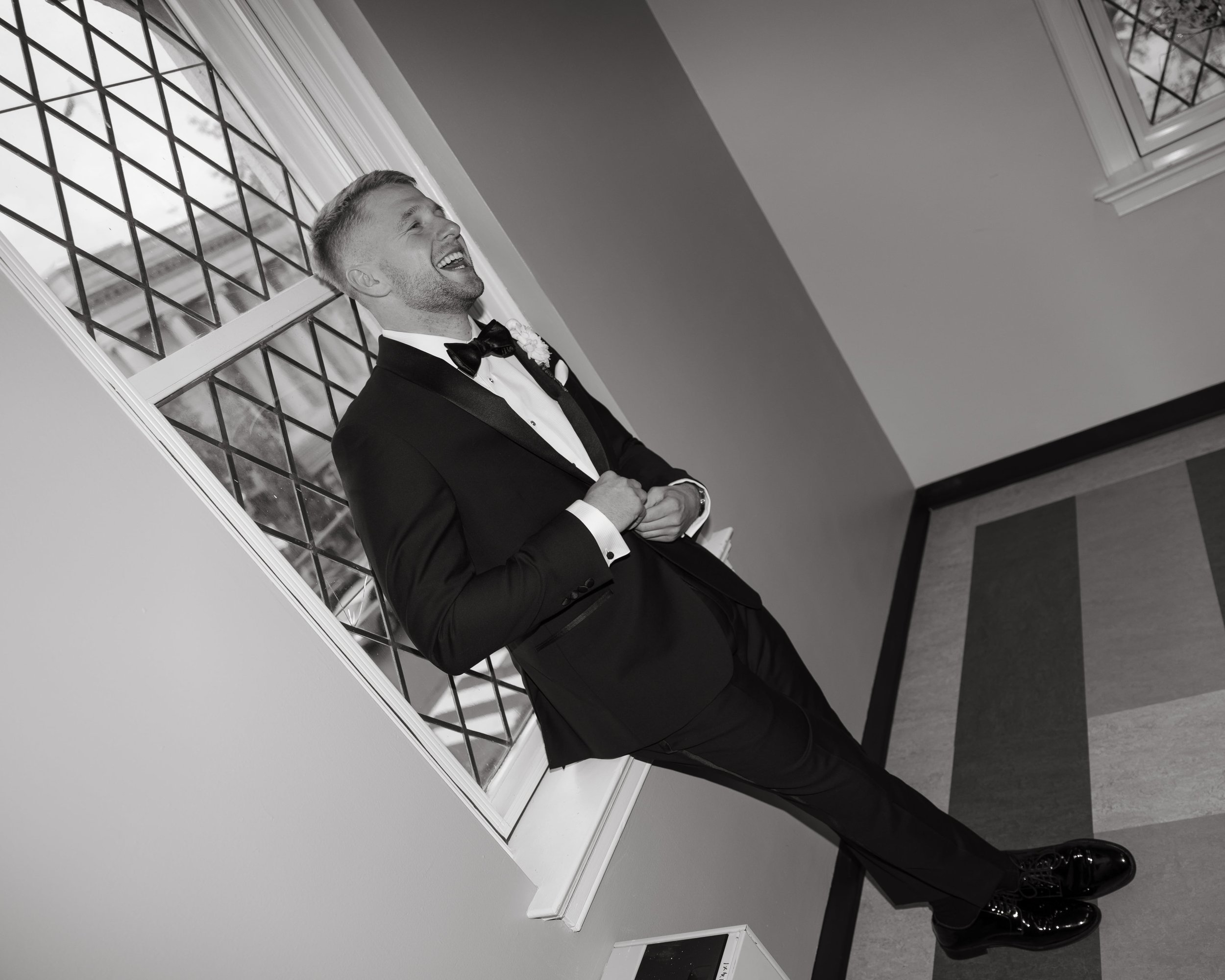 Black and white documentary wedding portrait of a groom leaning back against a tall window with diamond-pane glass at a historic DC church venue. He is dressed in a tailored black tuxedo with satin lapels, smiling and adjusting his jacket in a candid