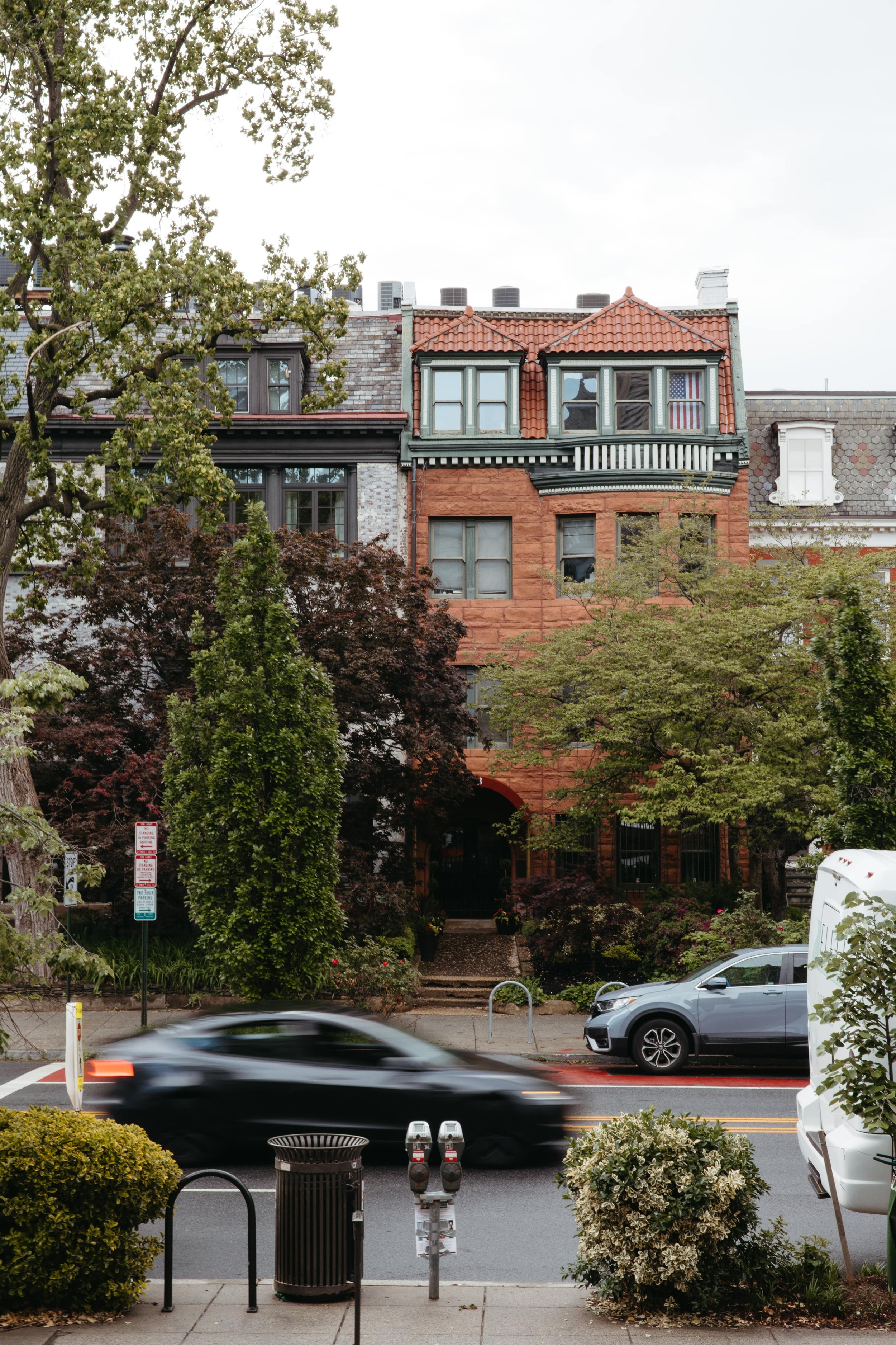 Photograph of a historic red brick row house with a green-trimmed roofline and American flag in the window, located on a tree-lined street in Washington DC. The foreground shows motion blur of a black car driving past, along with metered parking spac