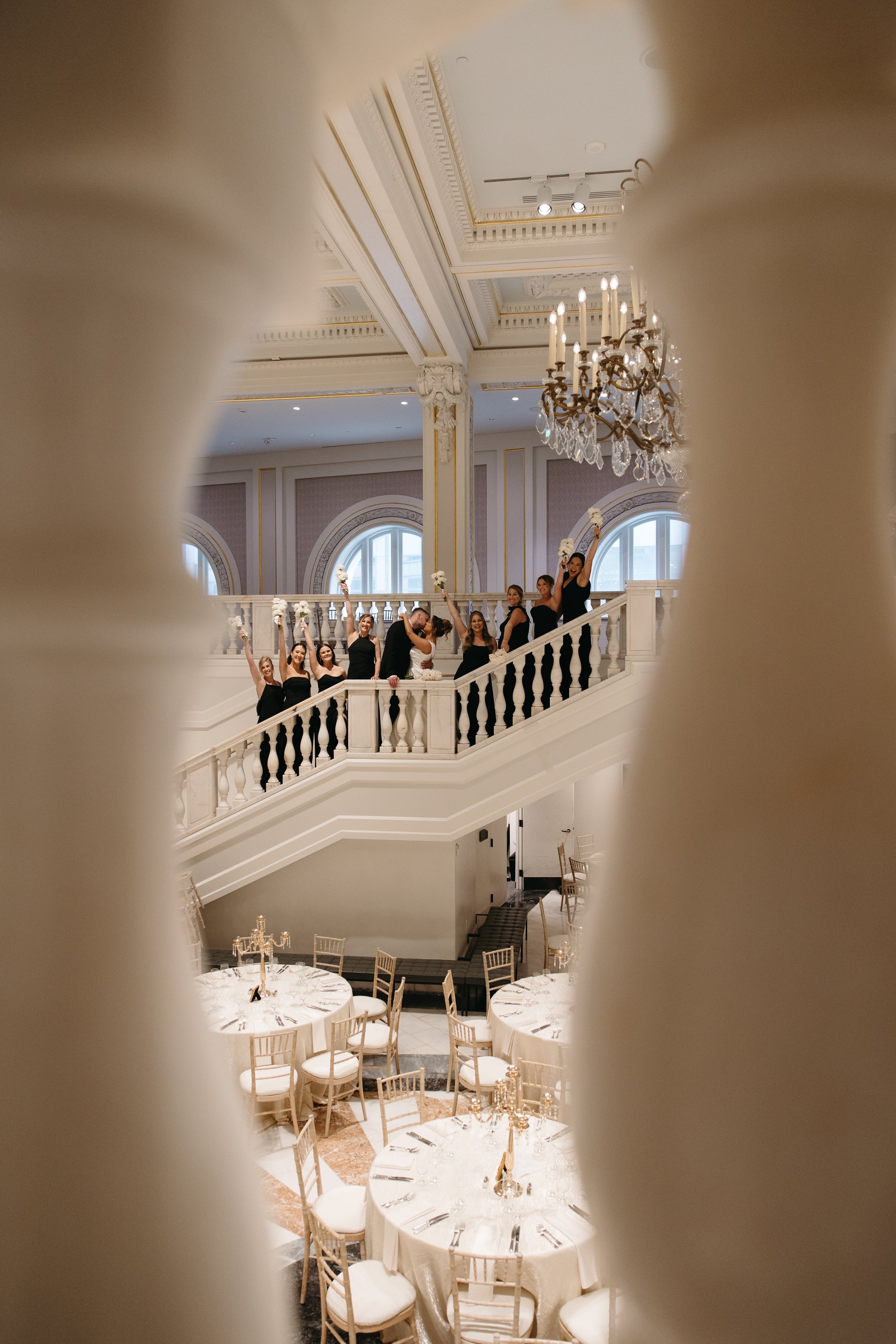 Creative wedding photography of bride and groom kissing on a grand staircase surrounded by bridesmaids holding bouquets in a luxurious ballroom with chandeliers and arched windows. Captured from a creative perspective through a balcony railing. Docum
