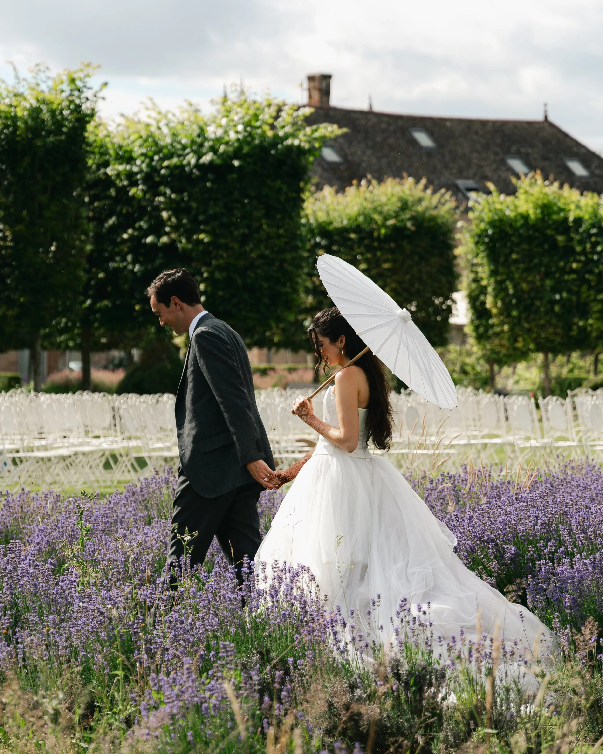 A bride in a tulle gown carries a white parasol as she walks hand in hand with her groom through a lavender field at Chateau de Varennes, Burgundy, France. Photographed by Kathleen Baker Photography, DC, NYC, and destination wedding photographer.