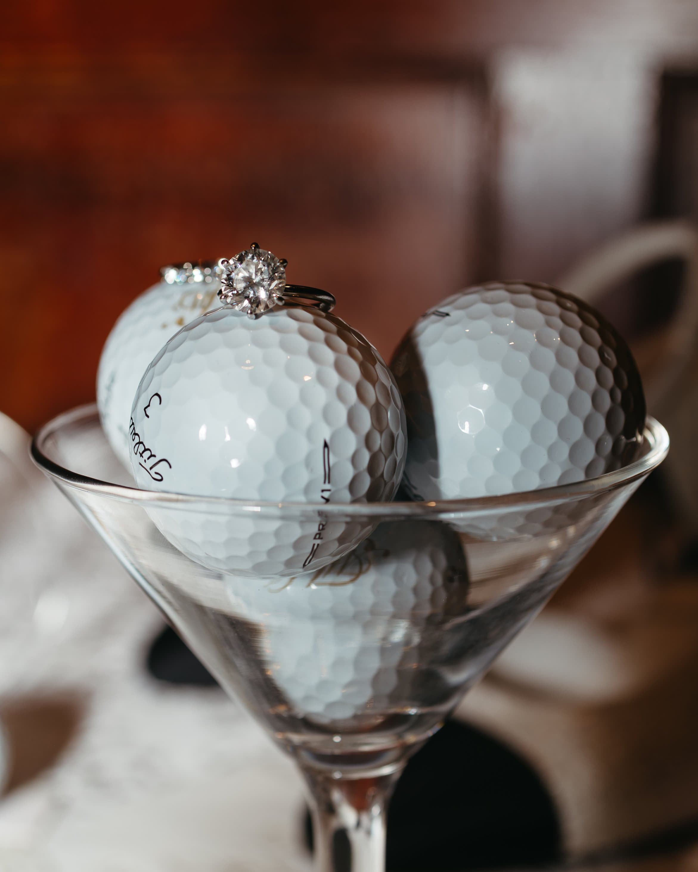 A creative display of a 3-carat solitaire diamond engagement ring delicately placed atop Titleist golf balls inside a martini glass at the prestigious Harvard Club in New York City. The glossy surface of the golf balls reflects the warm, elegant ligh