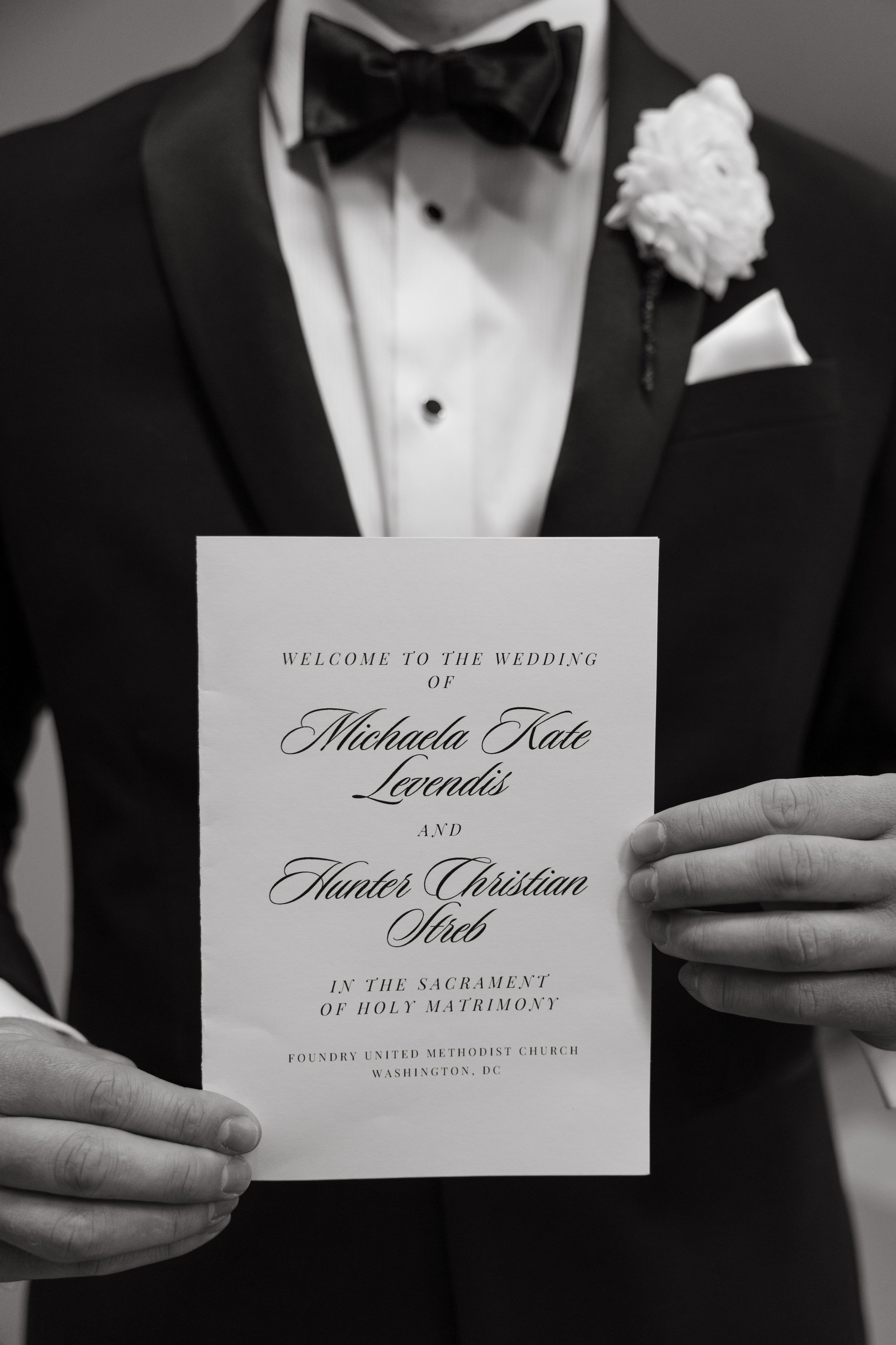 The groom holds the wedding program at Foundry United Methodist Church, showcasing the elegant calligraphy and refined paper design. His black tuxedo and white boutonnière provide a timeless frame for the invitation. The image is a close-up that hono
