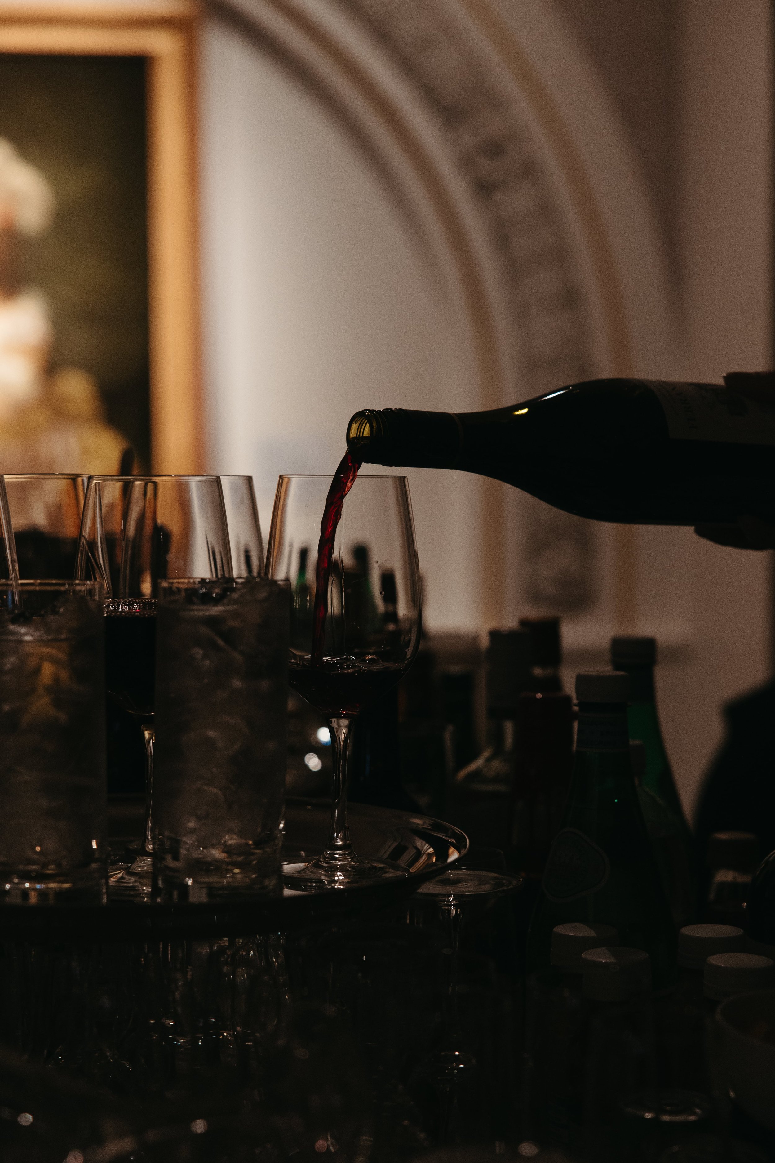 Moody editorial wedding reception photo of red wine being poured into stemmed glasses behind a bar setup with bottles of liquor and sparkling water. A soft-focus framed portrait and ornate arched molding are visible in the background. Captured at a l