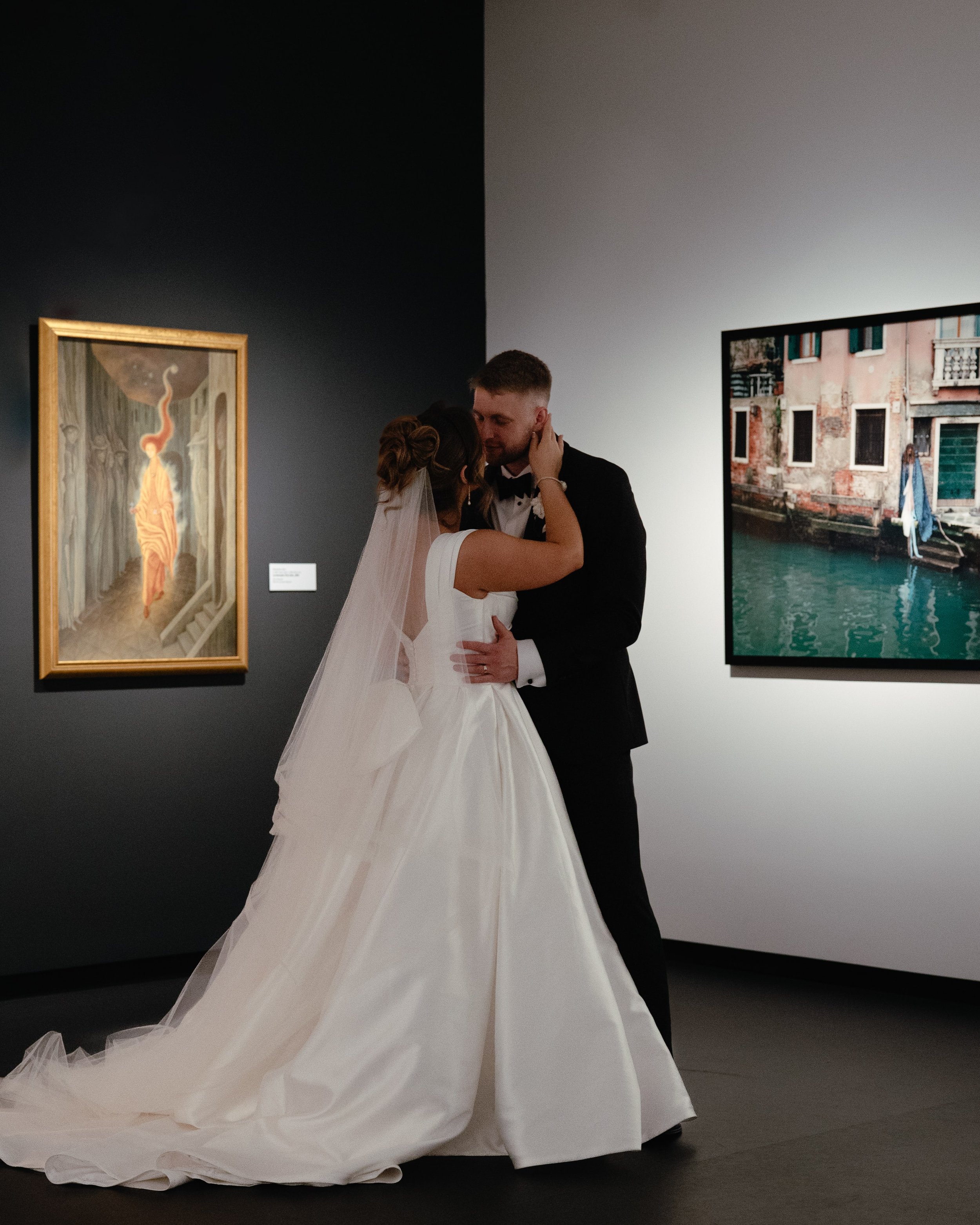 Editorial wedding portrait of bride and groom embracing in front of two large framed artworks in a contemporary art gallery. The bride wears a white gown with a long train and veil, and the groom wears a black tuxedo. Captured by Kathleen Baker Photo