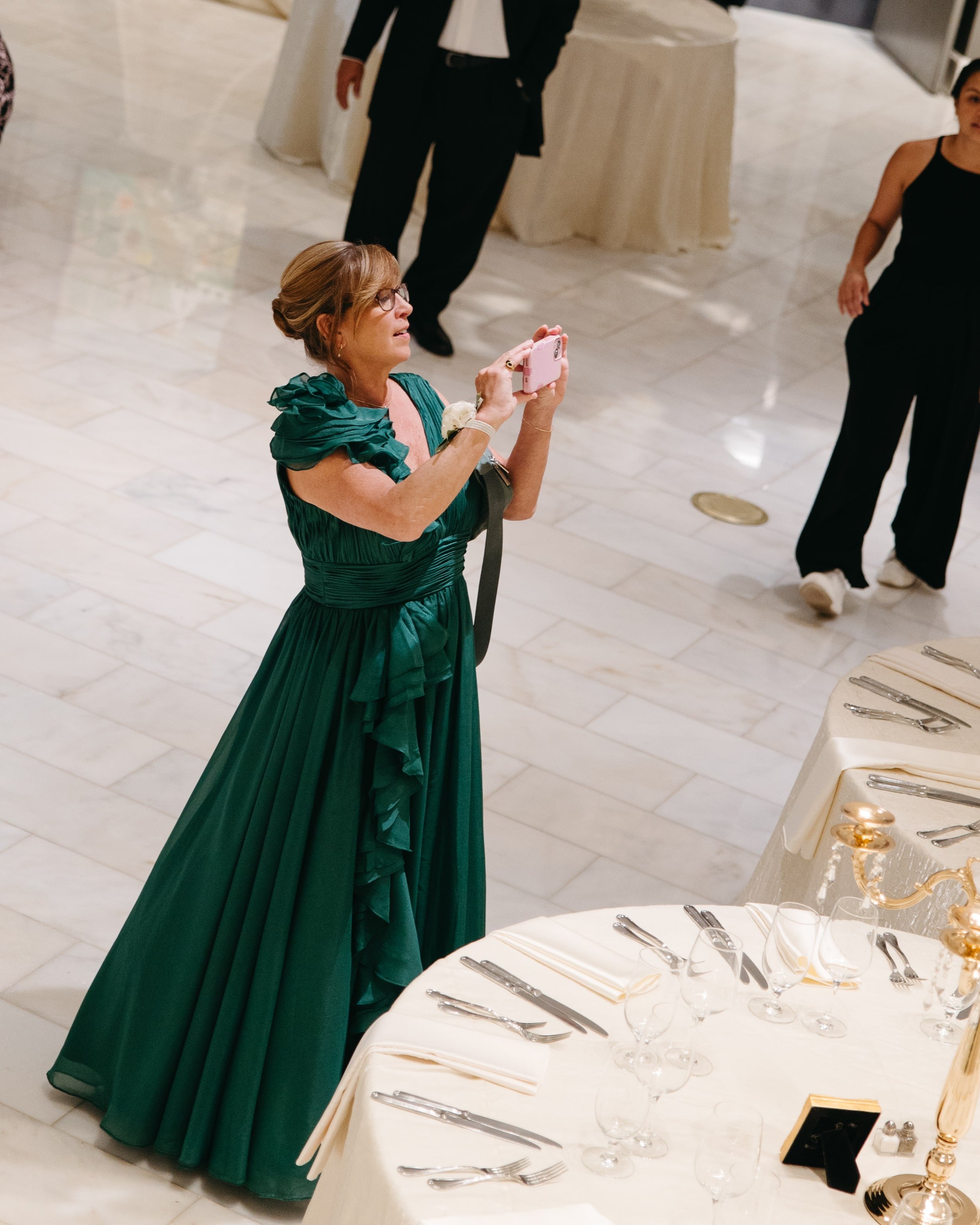 Editorial wedding reception photo of a woman in a green gown holding a pink smartphone taking pictures of the reception tables set with white linens, glassware, and gold candelabras in a marble ballroom. Documentary wedding photography by Kathleen Ba