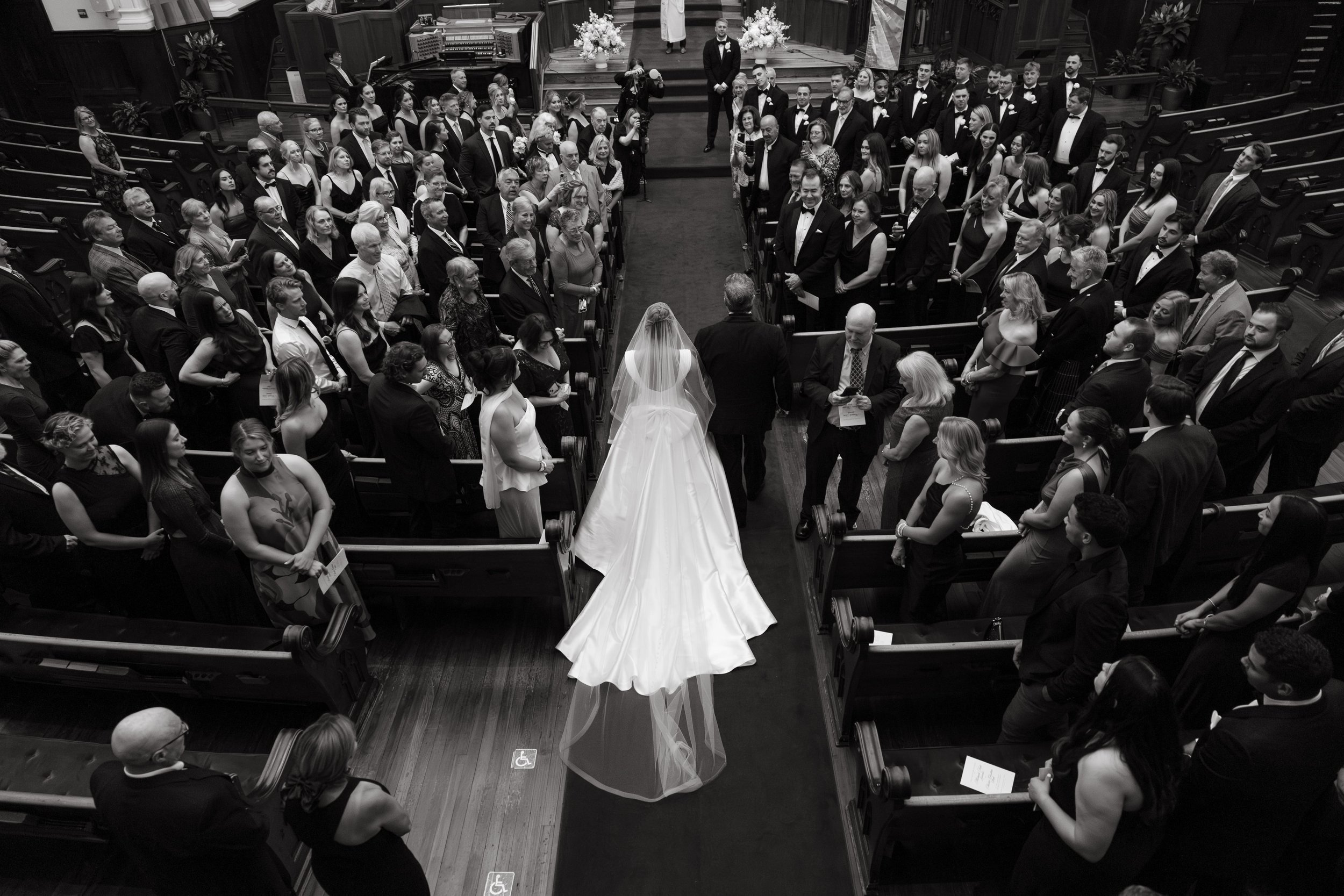 Black and white editorial wedding photography of a bride wearing a classic satin ballgown with a long train and veil, walking down the center aisle of a grand church in Washington DC. She is escorted by her father as guests in formal attire stand to