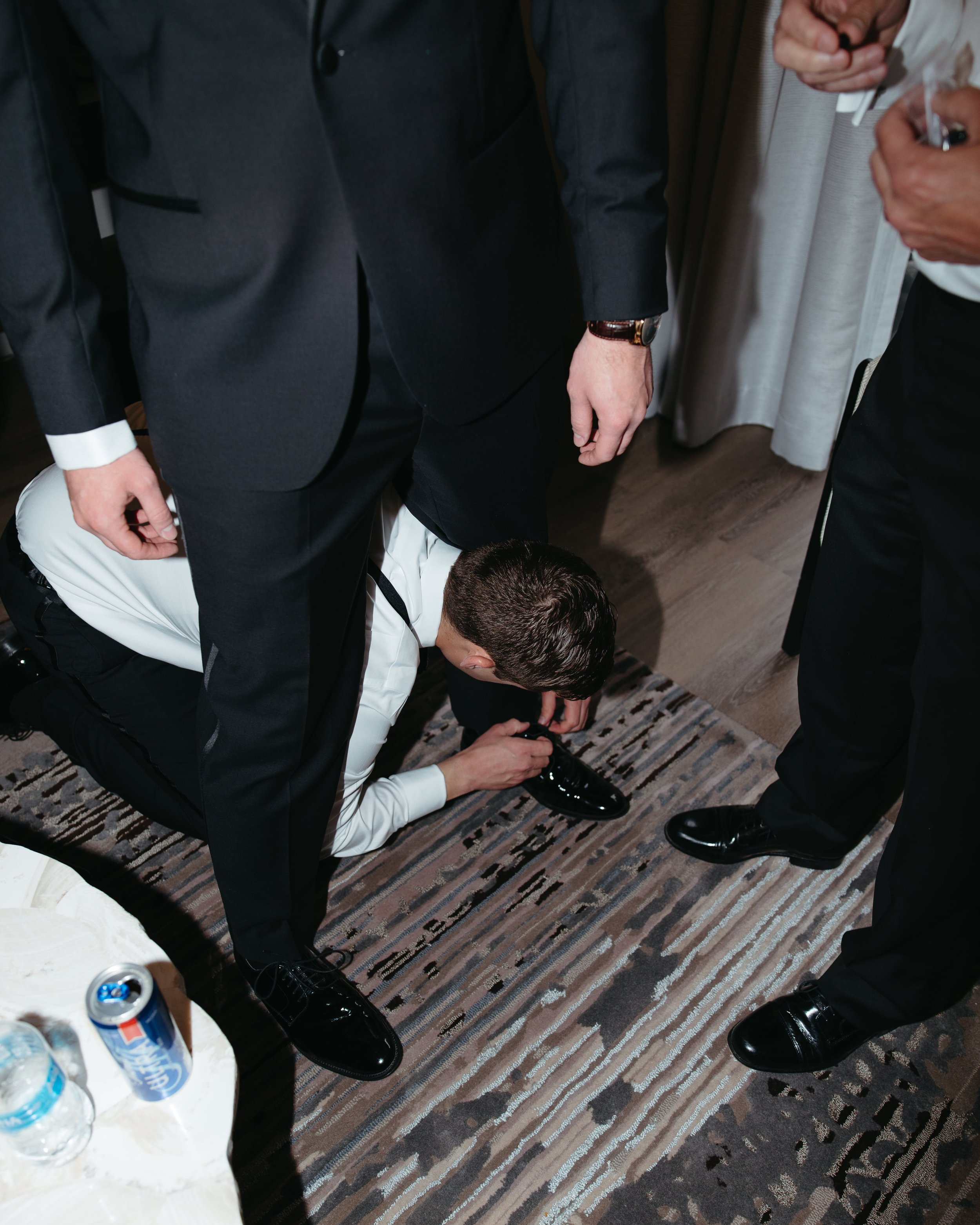 Documentary-style wedding photo of a groom’s suite in a modern DC hotel room, where a groomsman in a white dress shirt and black suspenders is crouched down tying the shiny black dress shoe of the groom wearing a tailored black tuxedo. The scene incl