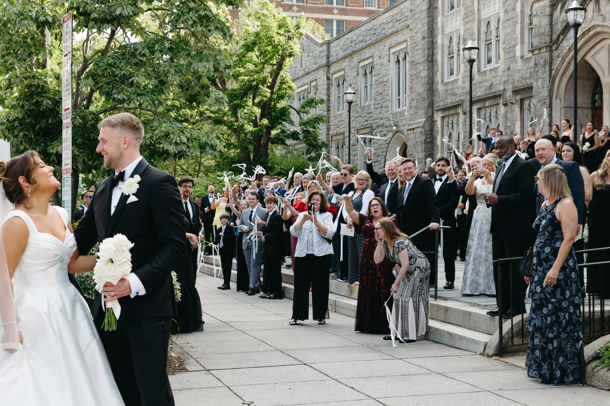 Documentary wedding photograph of a bride and groom walking hand in hand past cheering guests holding white ribbon wands outside a historic church in Washington DC. The bride wears an elegant off-shoulder gown and holds a bouquet of white flowers whi