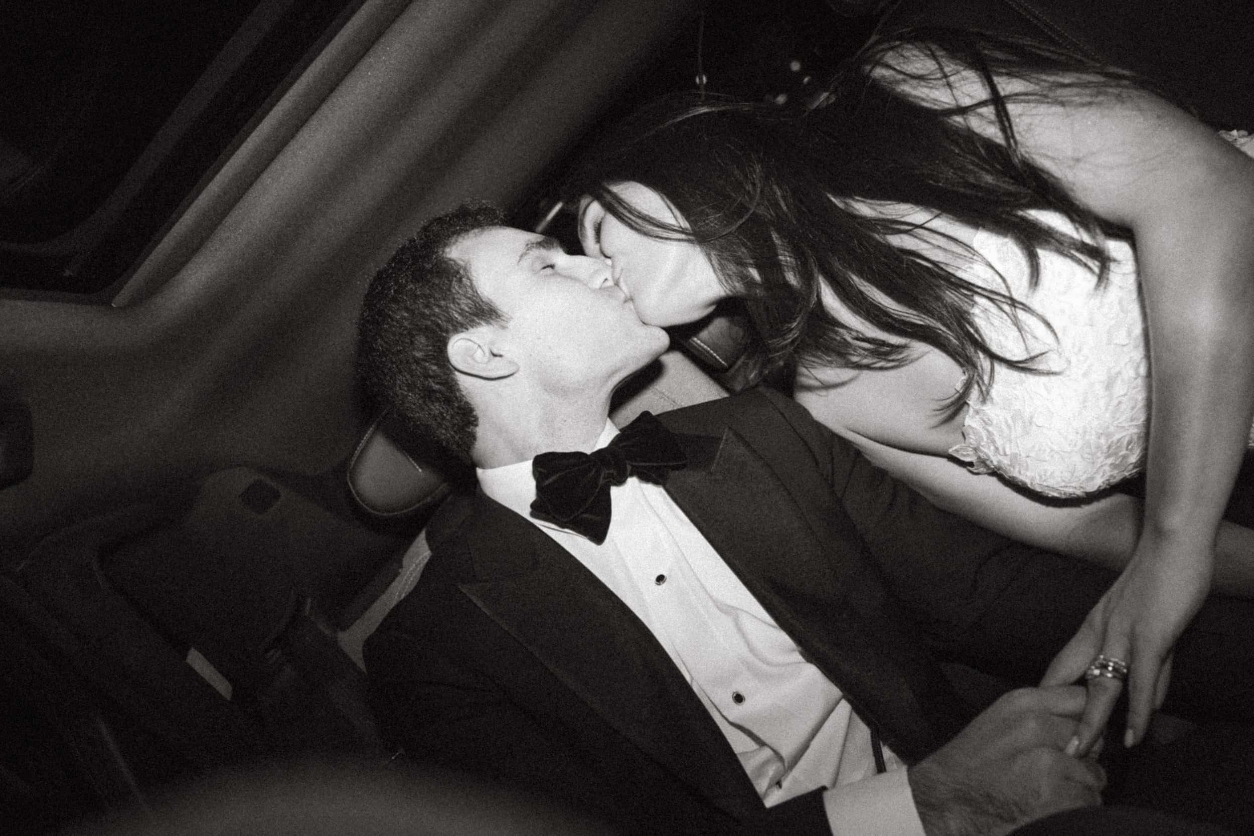 A black and white photograph captures a newlywed couple seated in the back of a luxury vehicle in New York City, sharing a kiss. The groom, wearing a classic black tuxedo with a crisp white dress shirt and an oversized black velvet bow tie, leans in