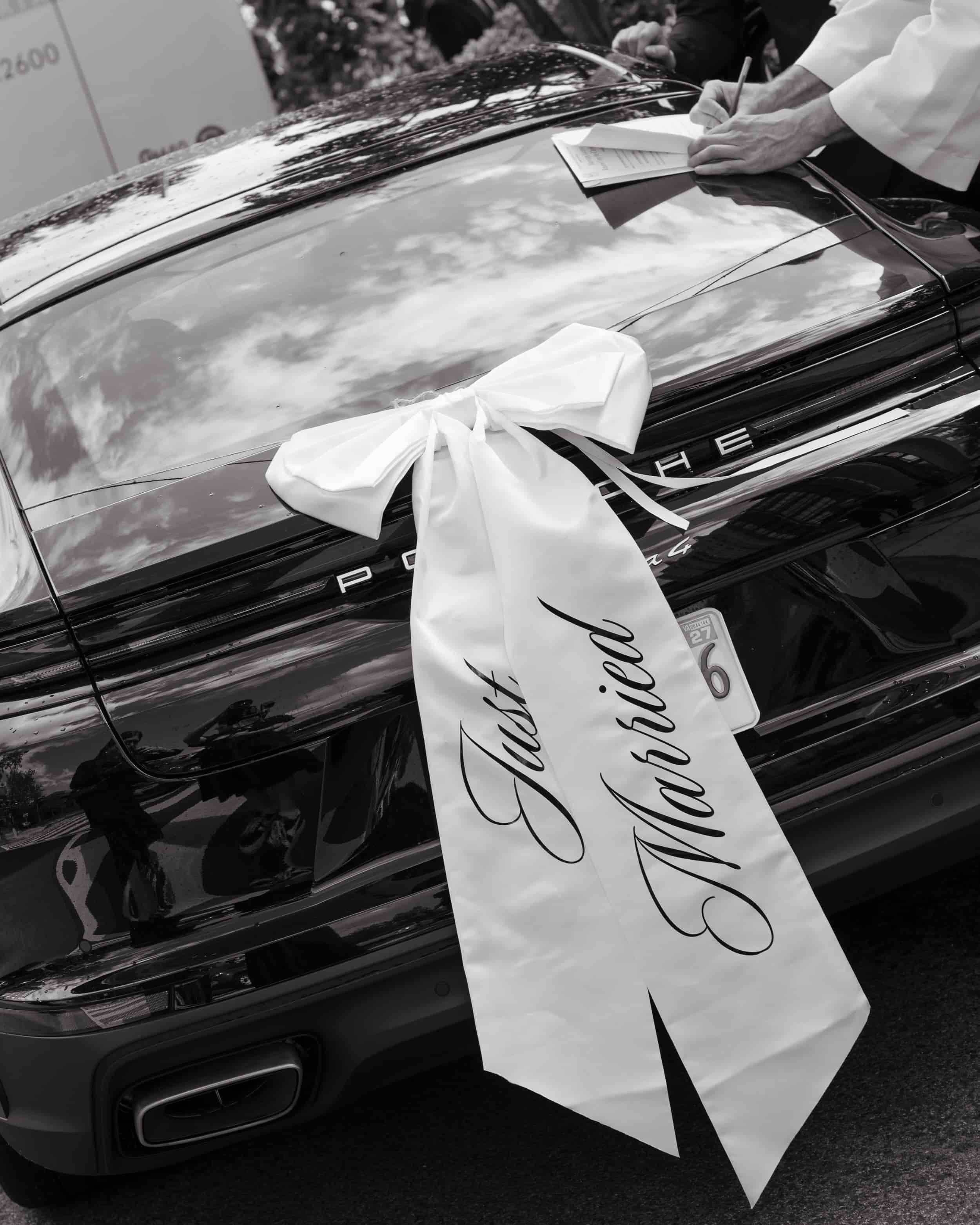 A monochrome image of a Porsche with a “Just Married” satin ribbon bow on the back, parked outside Foundry United Methodist Church in Washington, DC. A hand is seen signing the marriage license document atop the vehicle, symbolizing the final moments