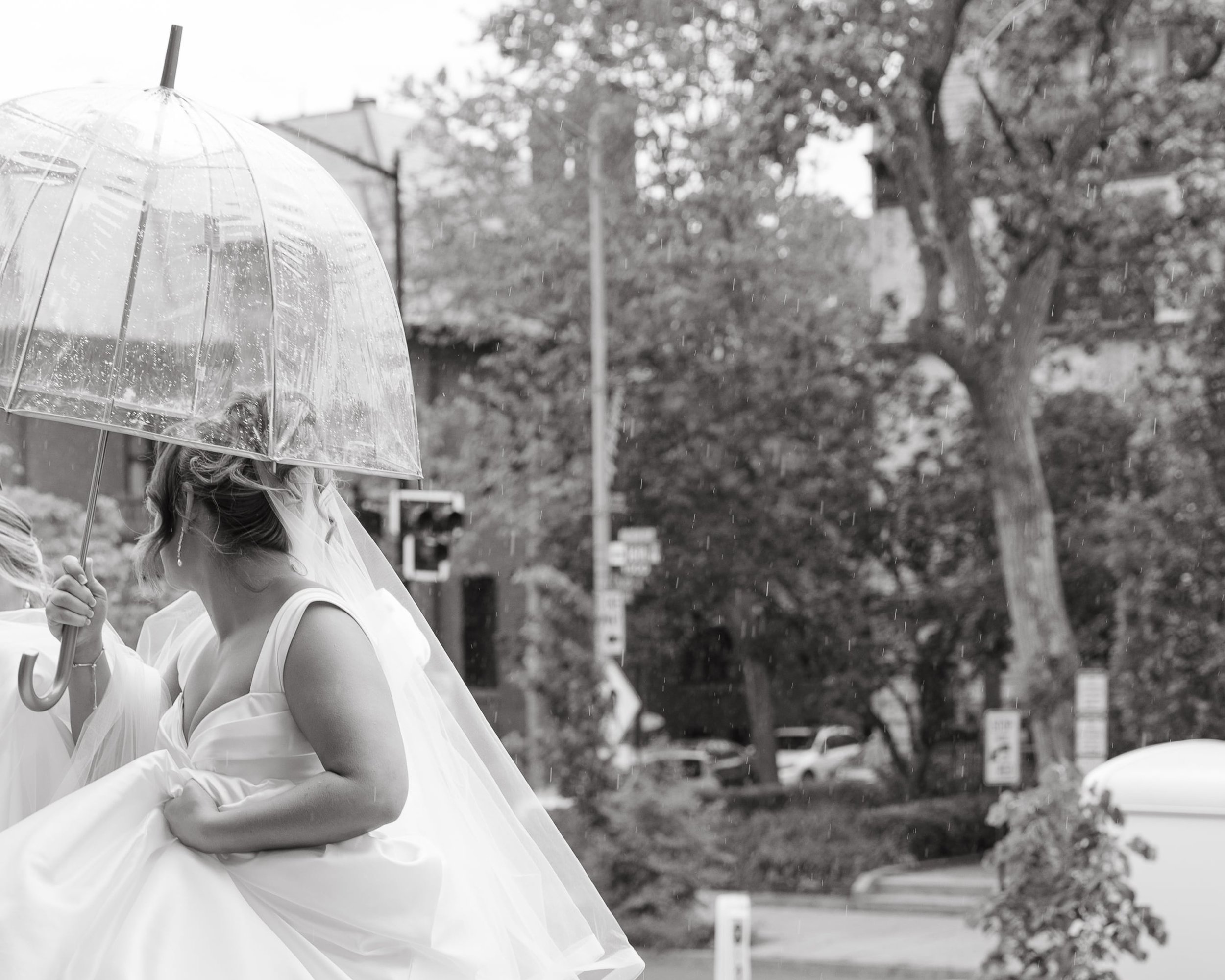 Black and white documentary wedding photograph of a bride in a sleeveless satin gown with a long flowing veil, holding the skirt of her dress in one hand and a clear umbrella overhead to shield from the rain. She walks up the stairs of a church in Wa