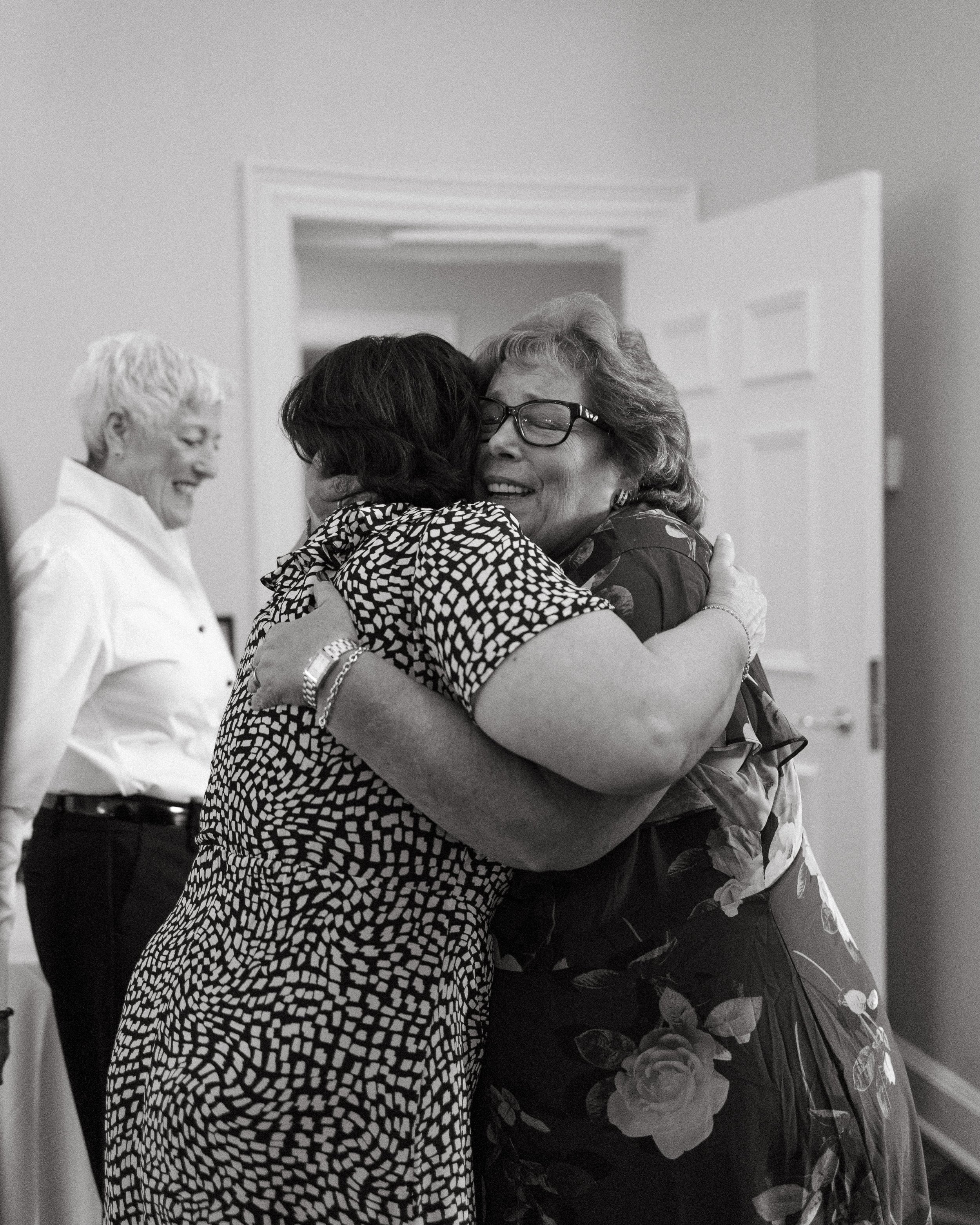 A black-and-white candid photo taken at Brantwyn Estate in Wilmington, Delaware during a rehearsal dinner. Two women embrace warmly, one wearing a black-and-white patterned dress and the other in a floral dress with large roses. Both are smiling with