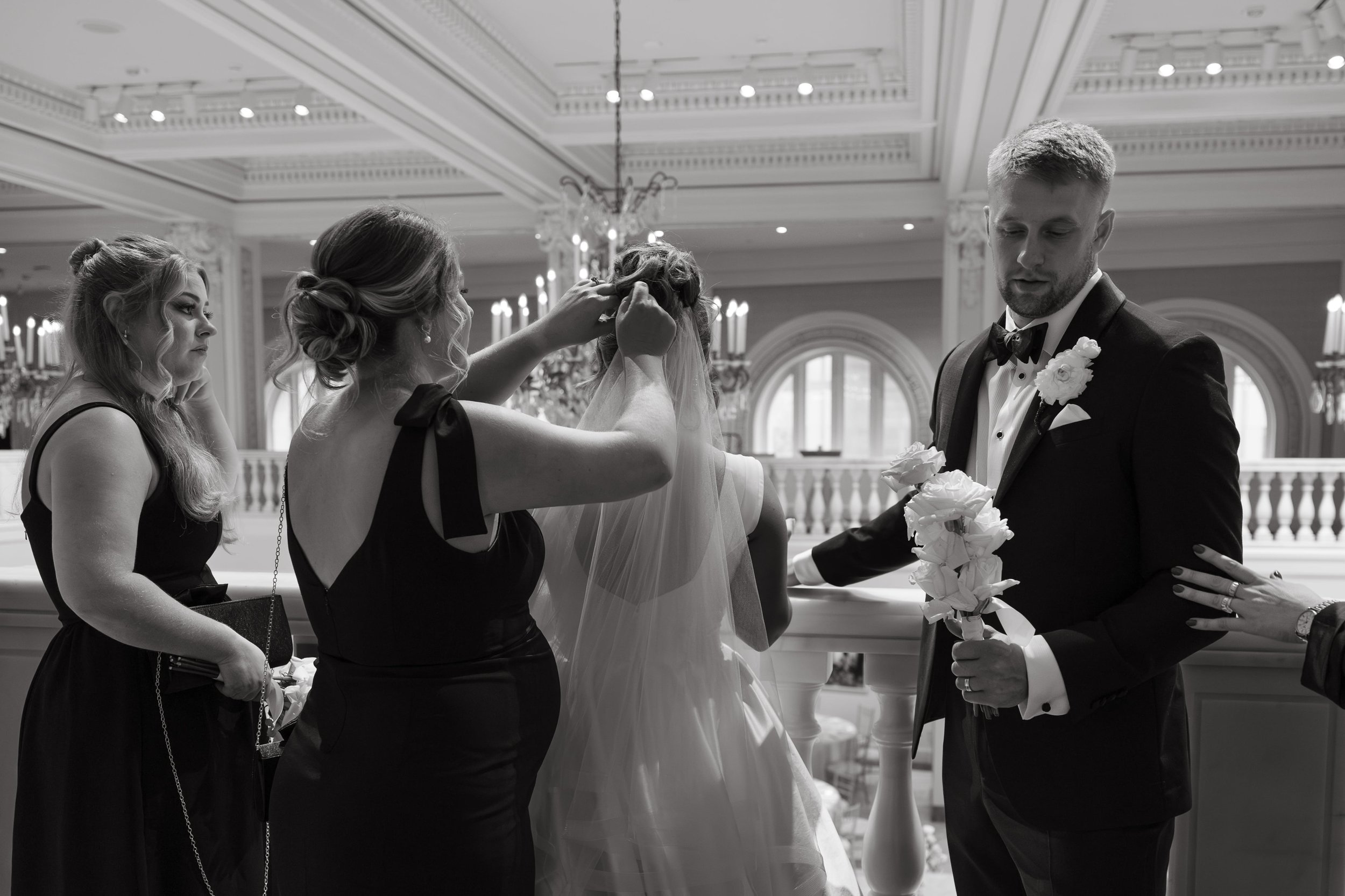 Black and white documentary wedding photo of the bride standing with her back to the camera as two bridesmaids adjust her veil in the Museum of Women in the Arts in DC with chandeliers and arched windows. The groom stands nearby holding a bouquet of