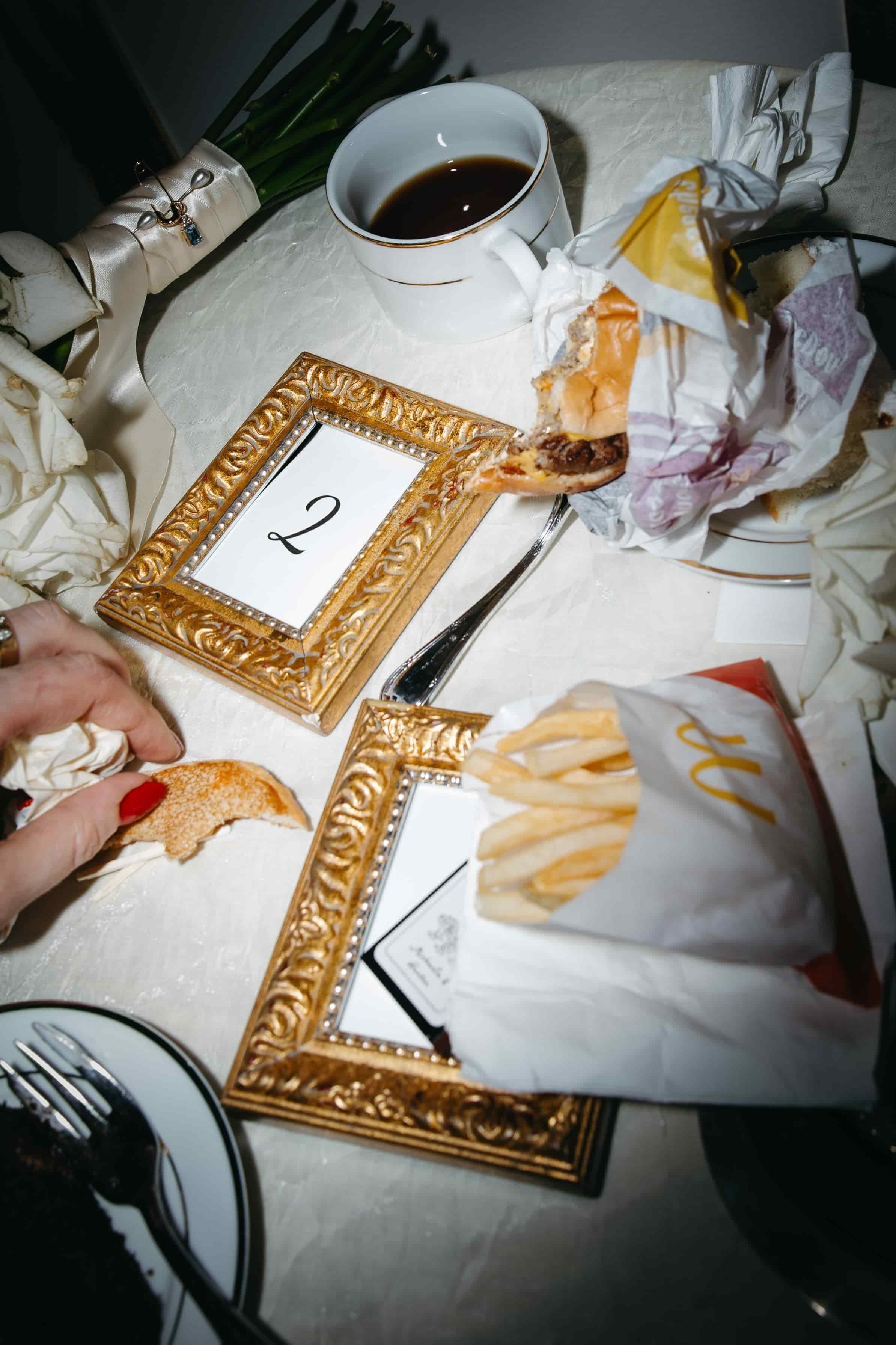A late night detail flatlay image with high-contract flash. There are table numbers laid down, matchboxes, a mug of coffee, and the bride's bouquet laid around. On top are a McDonald's burger and fries. Photographed during a luxury Washington DC wedd