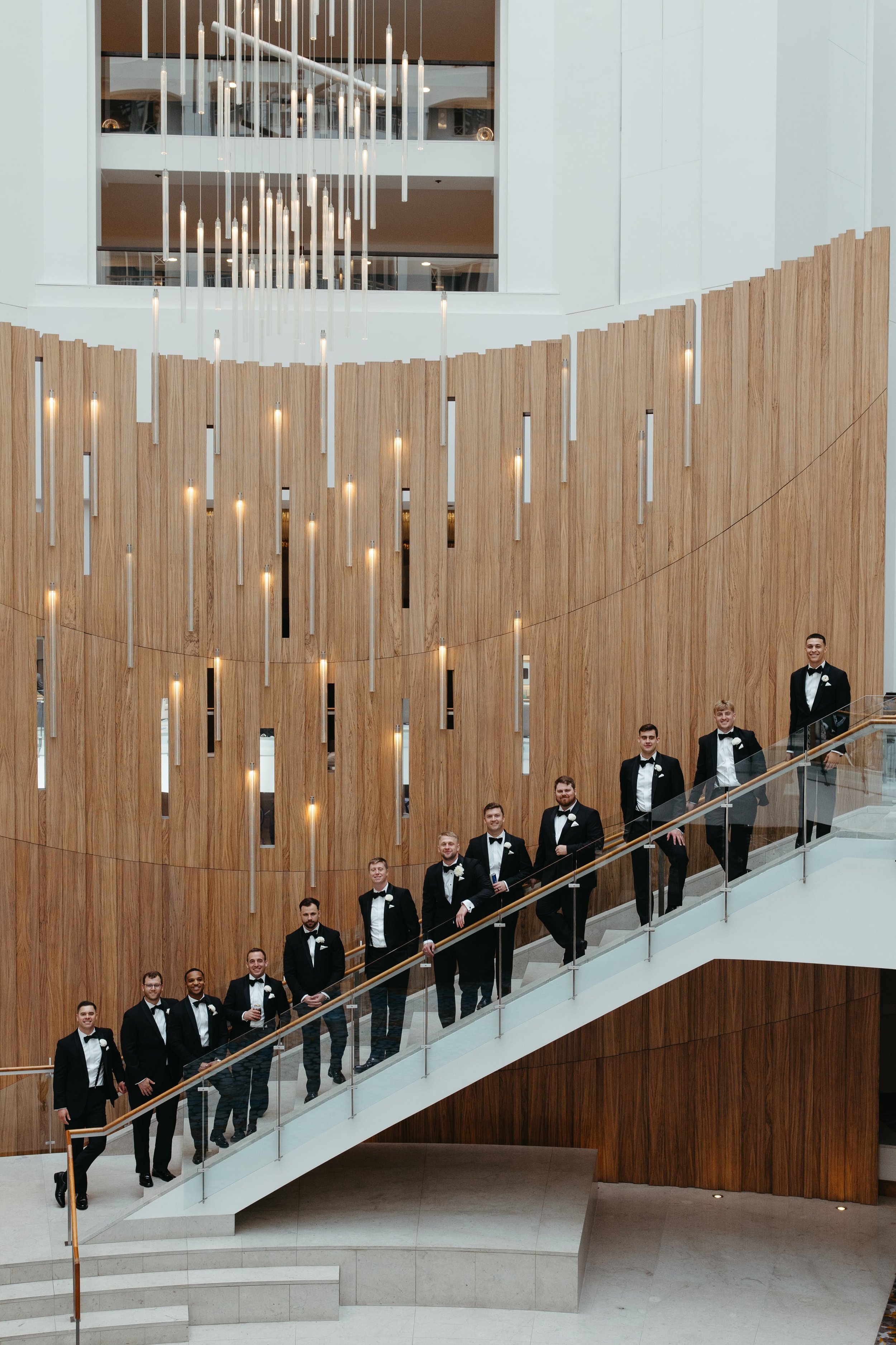 Editorial group portrait of eleven groomsmen dressed in black tuxedos and white boutonnieres, posed along a dramatic modern staircase with glass railings at a Washington DC Grand Hyatt. The backdrop features an architectural wall of vertical wooden s
