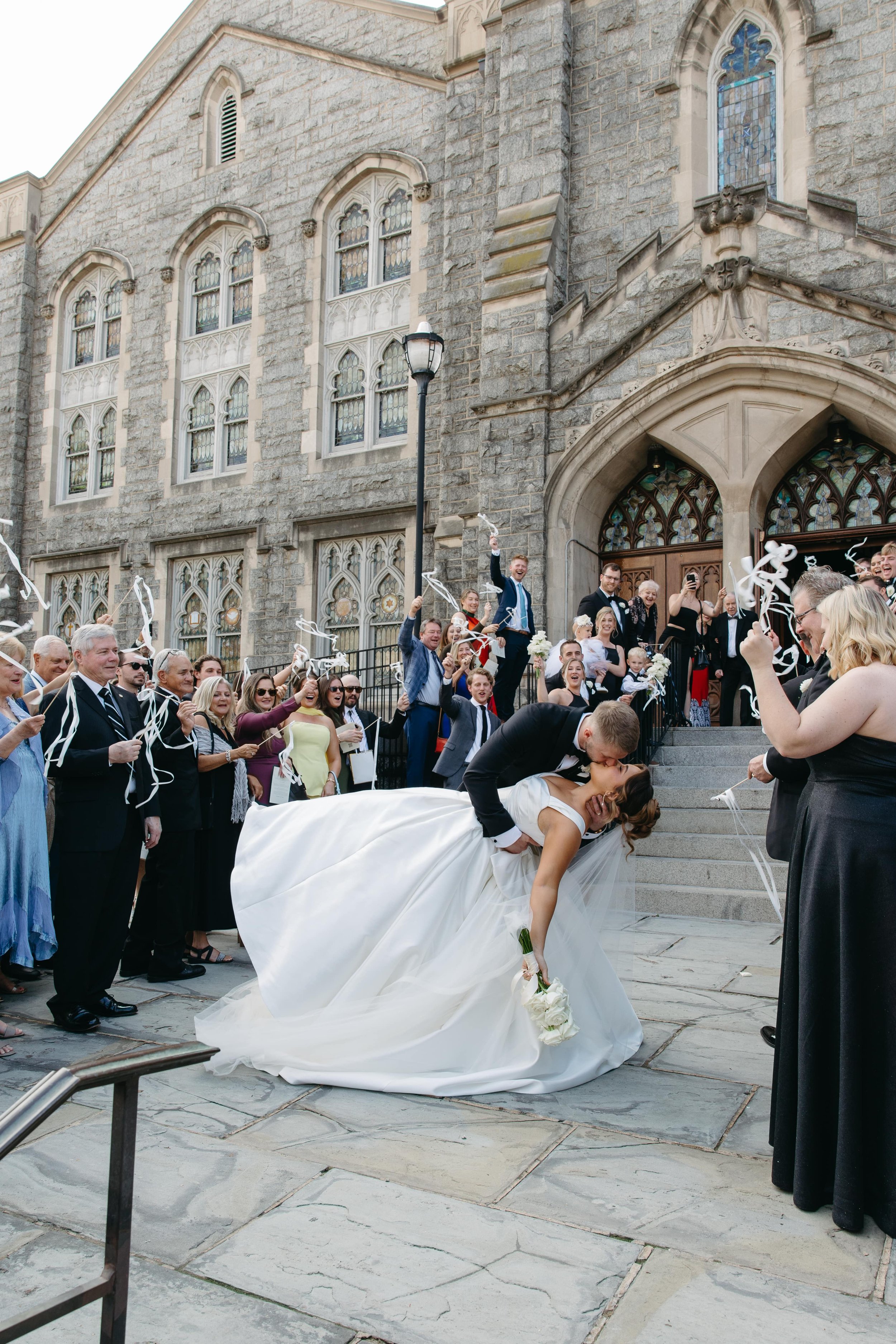Editorial wedding photo of a groom in a black tuxedo dipping and kissing the bride wearing a white satin gown while exiting a historic stone church with large arched doors and stained glass windows in Washington DC. Guests stand on either side waving