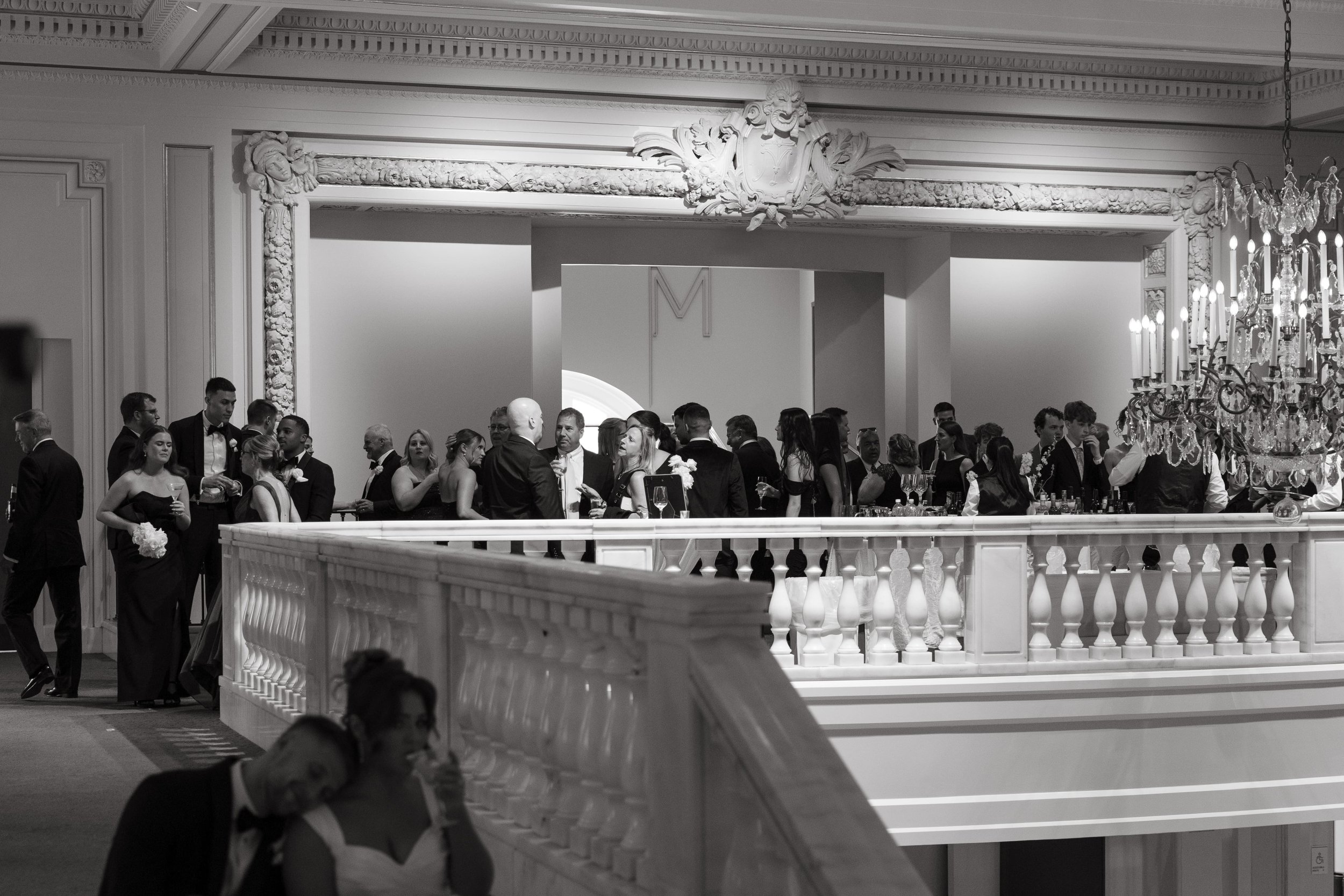 Black and white documentary wedding photo showing a large group of guests gathered at the upstairs balcony bar during cocktail hour in a grand ballroom venue. A chandelier and ornate plasterwork are in the background. In the foreground, the bride in
