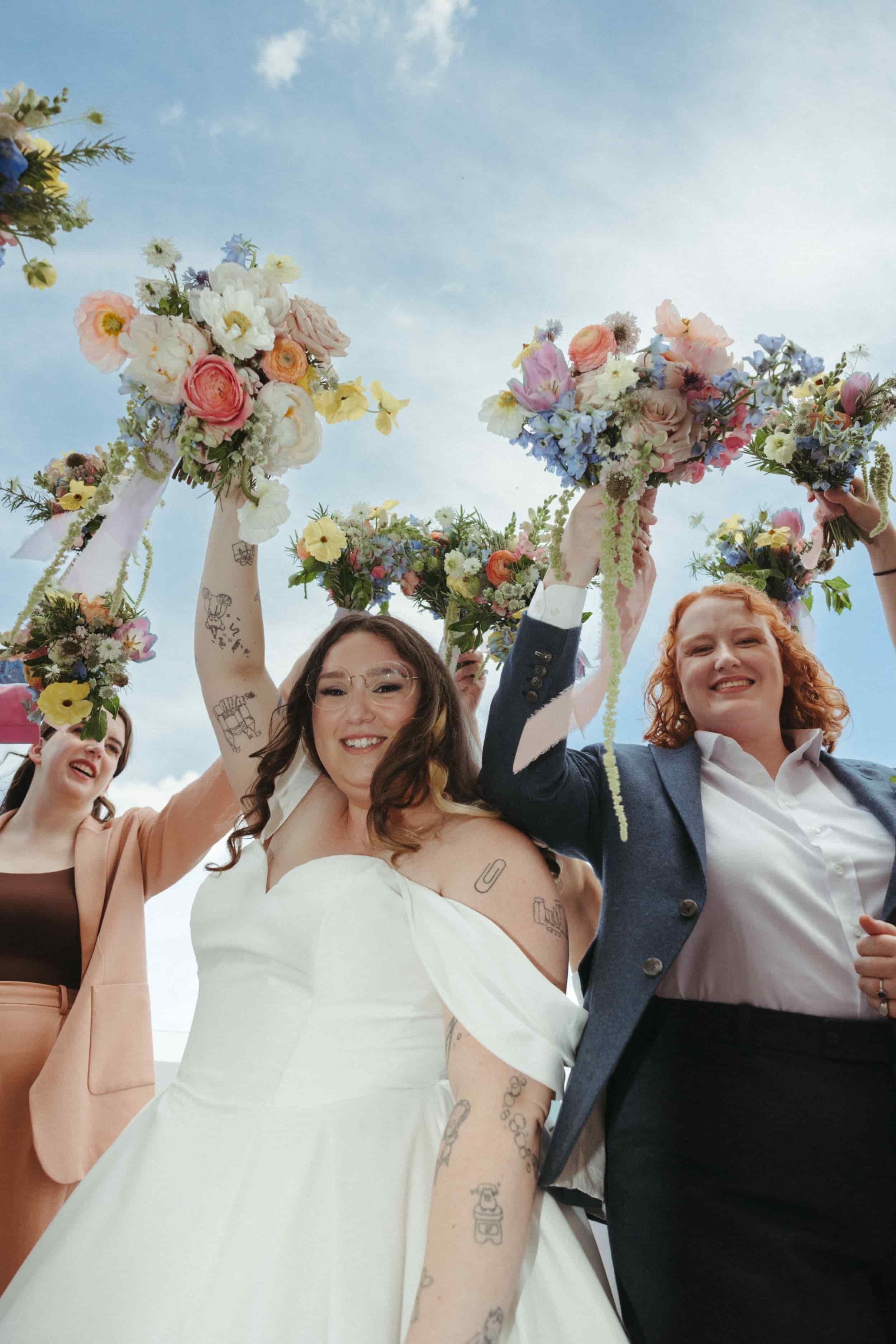Shot from below, a sapphic couple are surrounded by their wedding party, all holding their bouquets up to the sky. Bouquets feature tulips, ranunculus, and roses in pastel hues. Kathleen is a luxury wedding photographer offering documentary and edito