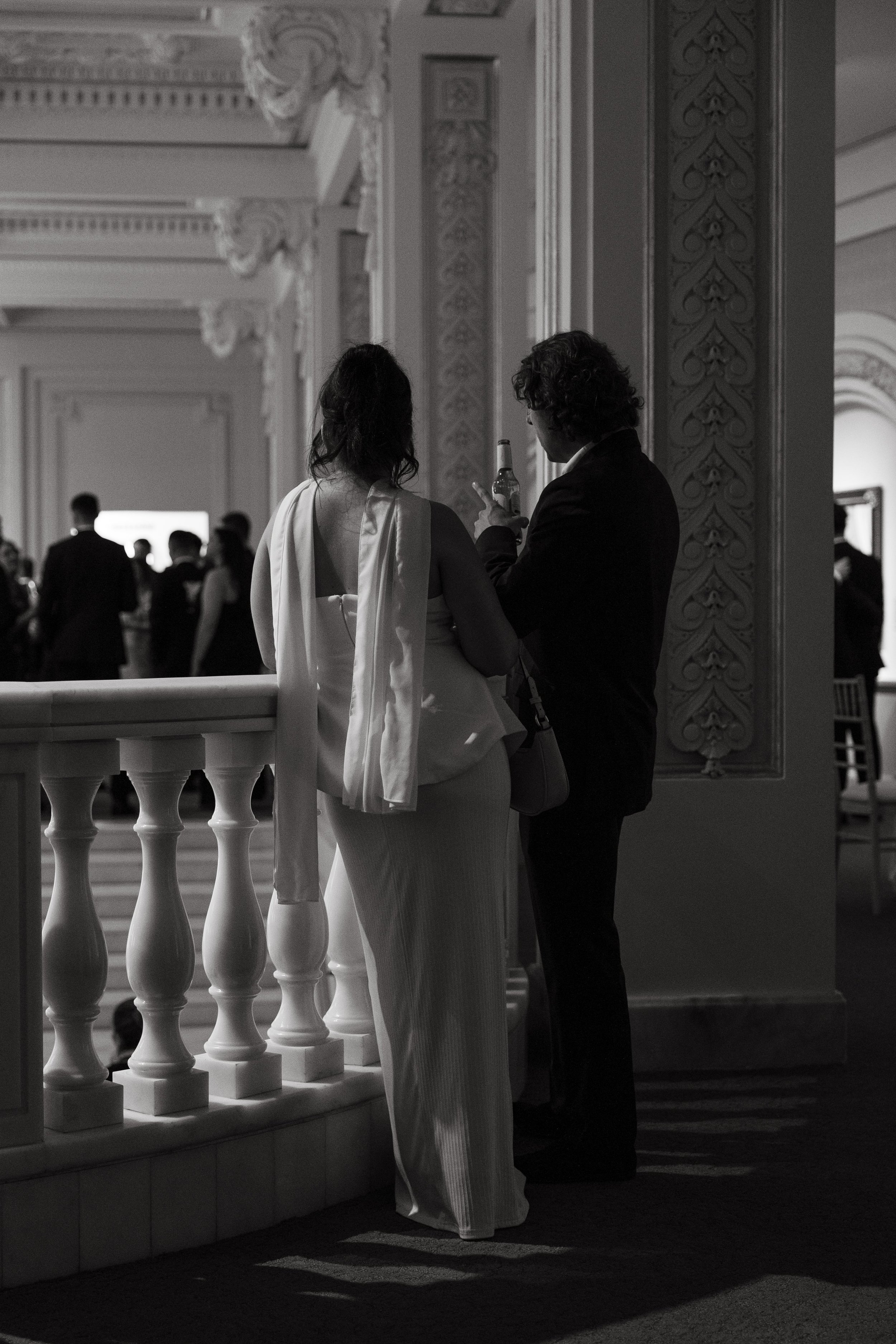 A candid black and white photograph taken by Kathleen Baker Photography, a DC and NYC wedding photographer, showing two wedding guests during cocktail hour at the National Museum of Women in the Arts. The man in a dark suit holds a beer bottle while