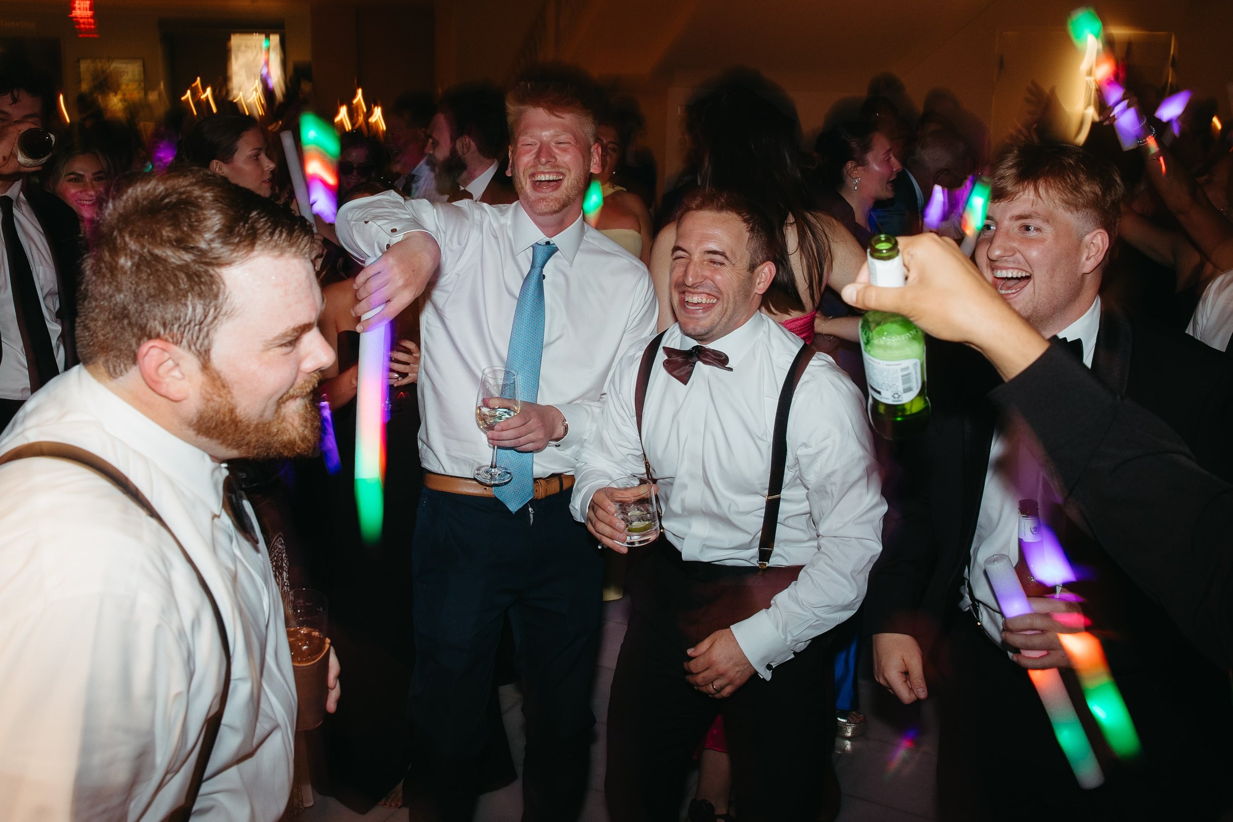 Dynamic documentary wedding reception image by Kathleen Baker Photography capturing groomsmen and guests partying with colorful LED foam sticks on the dance floor. Subtle shutter drag. The scene is filled with movement and laughter as men in white sh