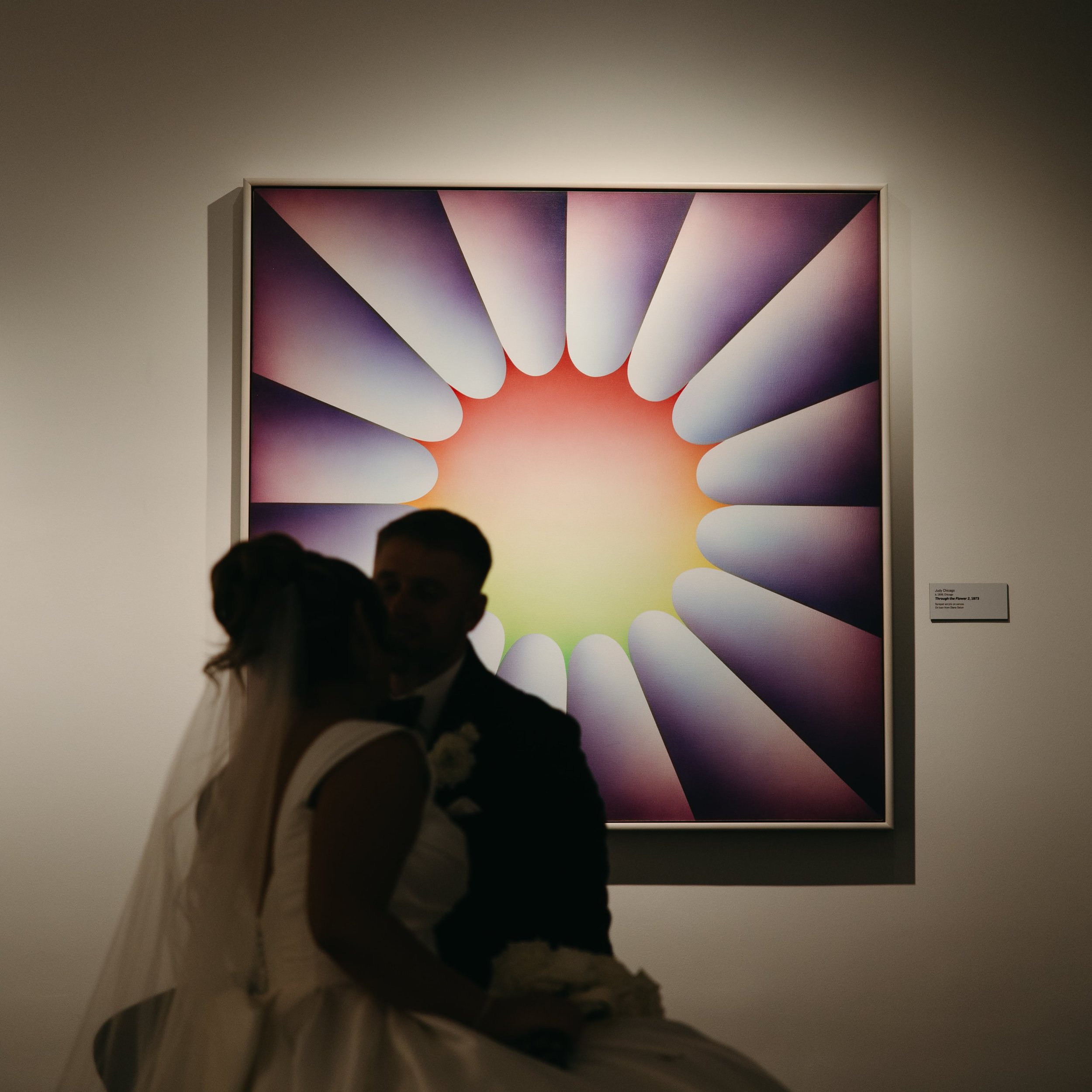 Artistic wedding photo of a bride and groom silhouetted in front of a large modern painting with a colorful starburst design in a gallery setting. The bride wears a satin gown and veil while the groom is in a black tuxedo. Editorial documentary weddi