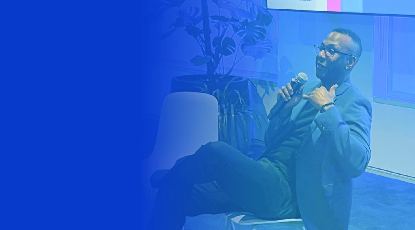 Thomas Shillingford UX Thought leader, a blue blazer, seated and holding a microphone, speaking in a room with plants in the background, overlaid with a blue gradient.