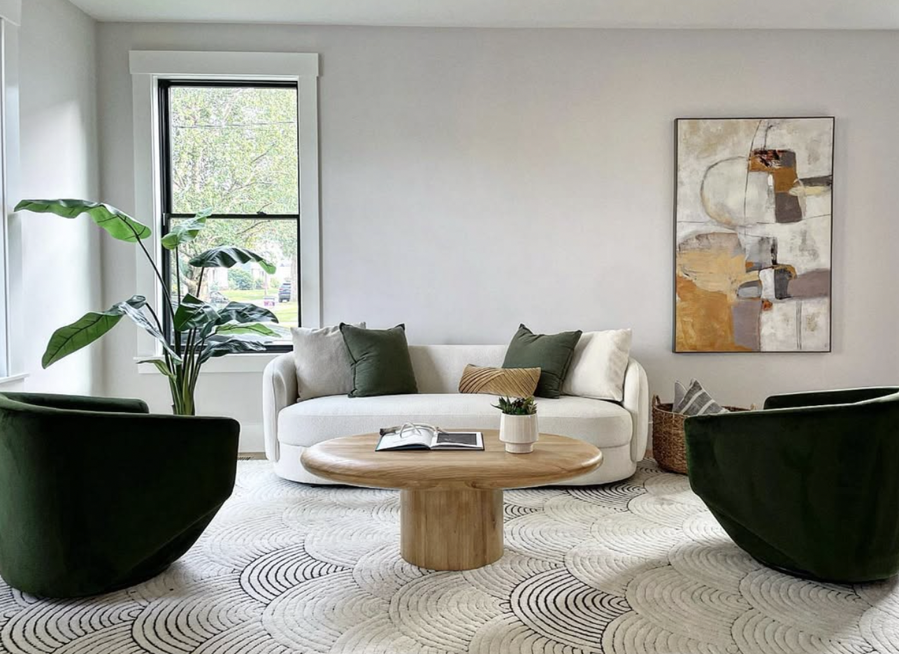 Modern living room with a white sofa, green and beige pillows, a wooden coffee table with a plant and open magazine, dark green armchairs, a large plant near the window, abstract wall art, and a patterned rug.