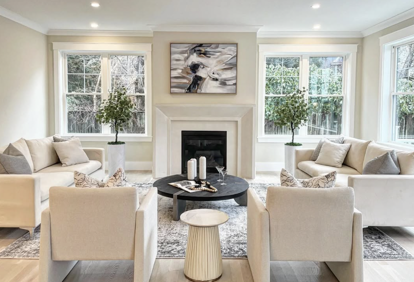 Bright living room with white sofas, a black coffee table, a fireplace, and large windows with trees outside.