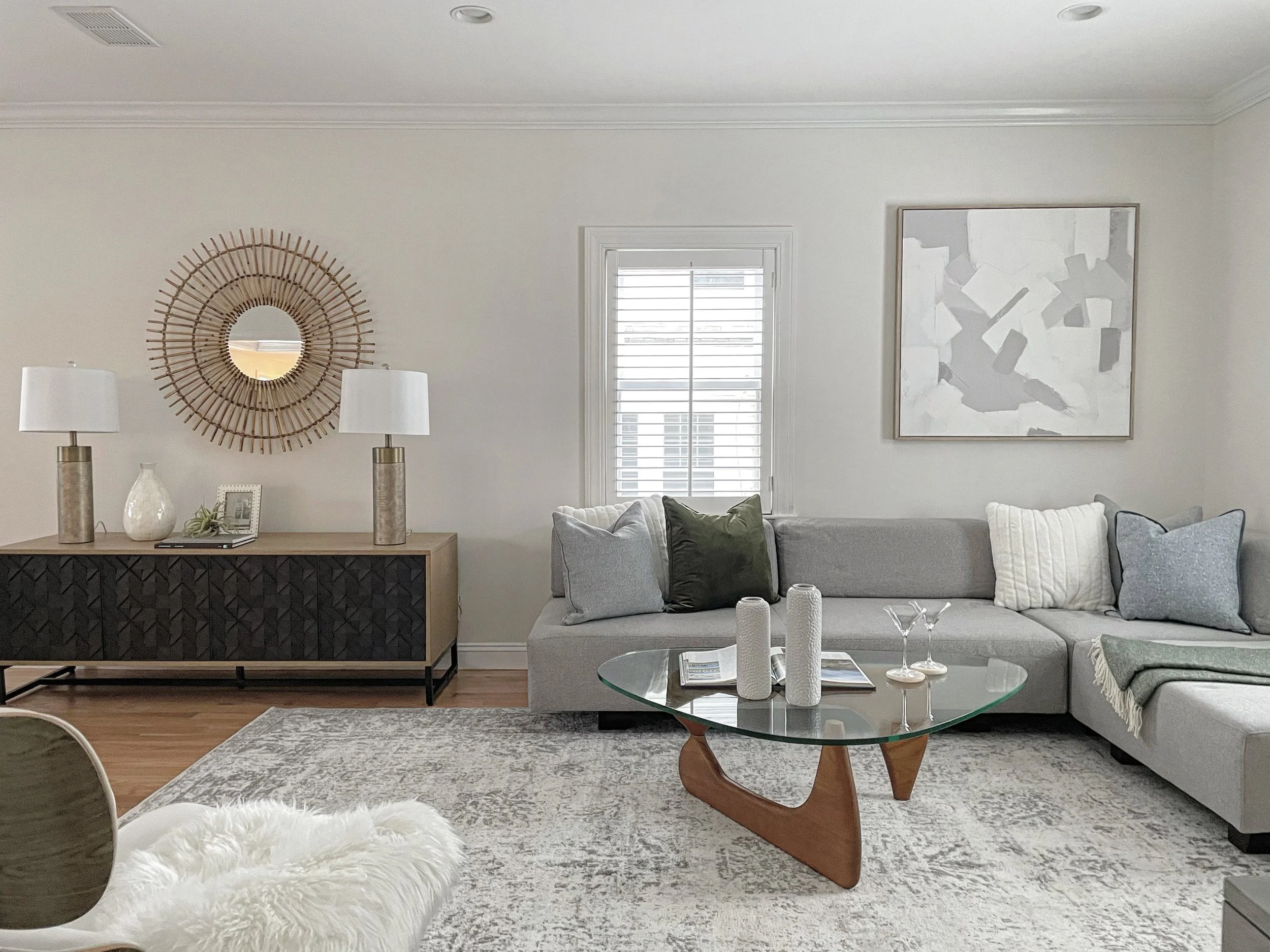 Modern living room with gray sofa, white pillows, abstract artwork, wooden coffee table with white decorative items, glass top, porcelain vase, framed photo, two white table lamps, circular mirror with wooden frame, window with white blinds, white walls, ceiling with recessed lighting, area rug, and hardwood flooring.