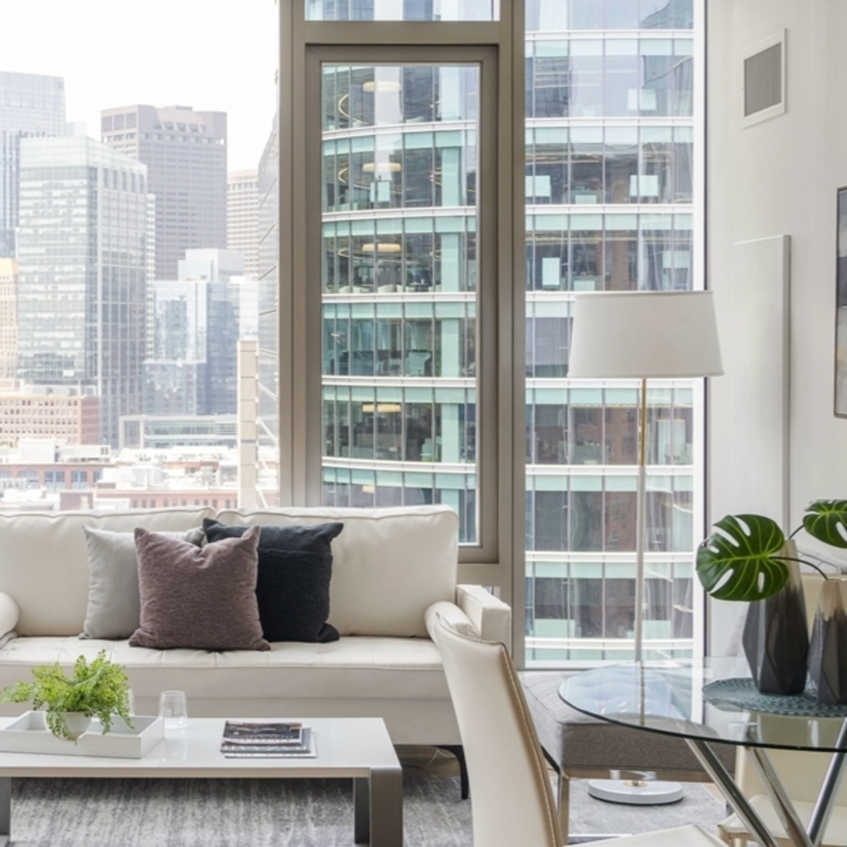 Interior living room with large floor-to-ceiling windows showing city skyscrapers, a white sofa with gray and black pillows, a small white coffee table with magazines and a plant, a glass side table with a large potted plant and a white lampshade.