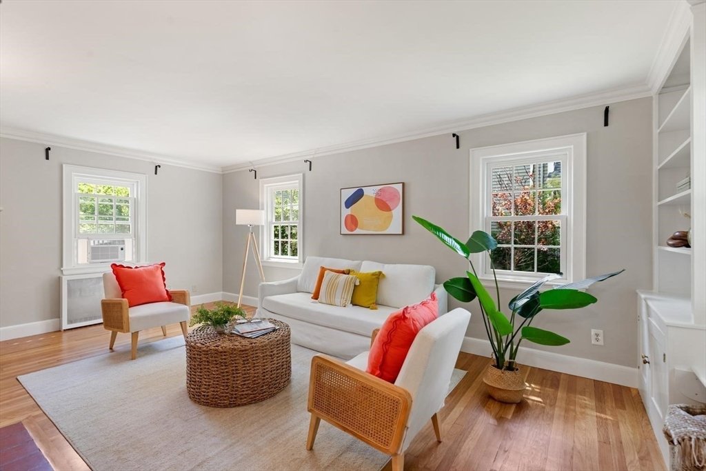 A white sofa with bright pillows and bright art. Two rattan armchairs and a light wood tripod lamp.