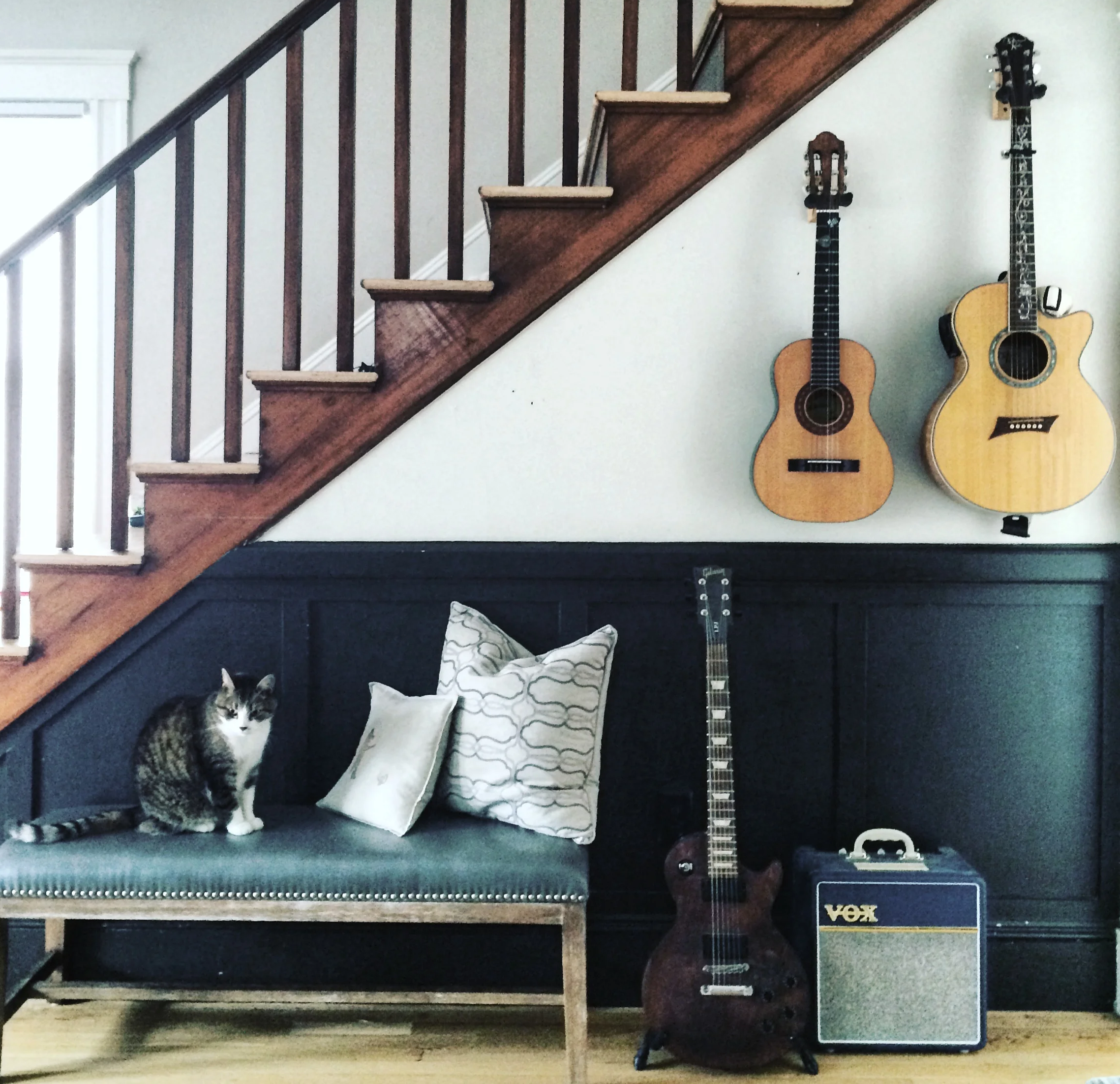 A wall with two acoustic guitars hanging next to a staircase, a small cat sitting on the floor in front of a pillow, and a VOX amplifier on the floor.