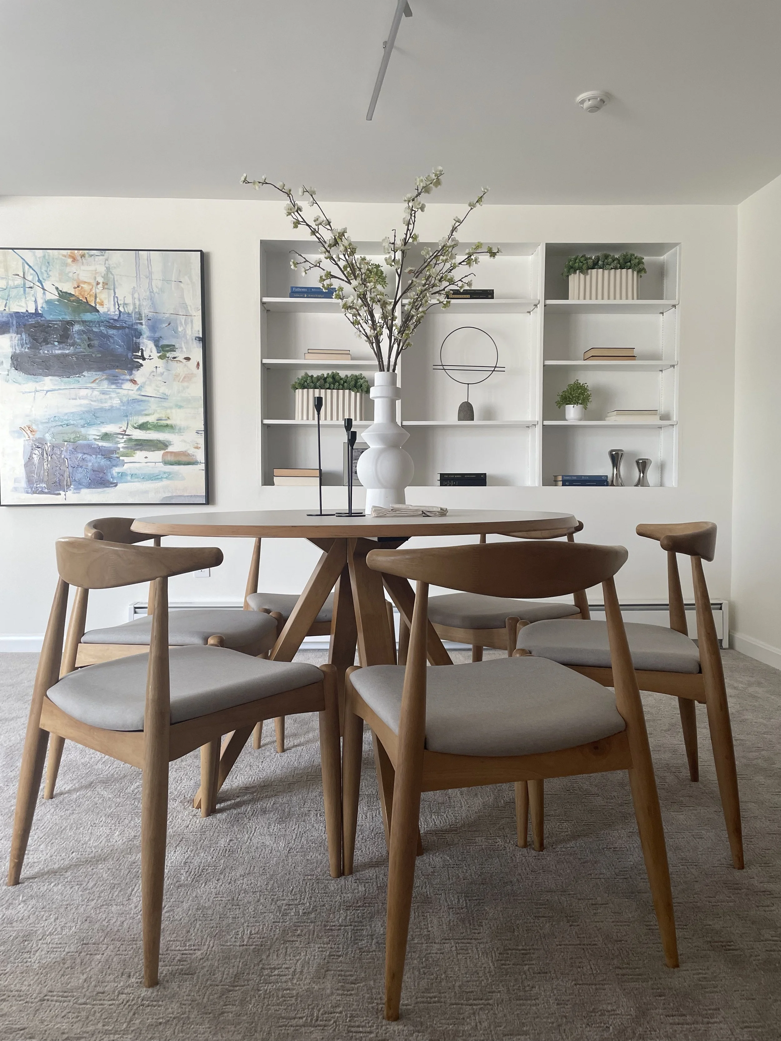 A round white and wood dining table with midcentury base and light wood upholstered wishbone chairs. There is a large white vase as a centerpiece with greens. Modern art in the background.