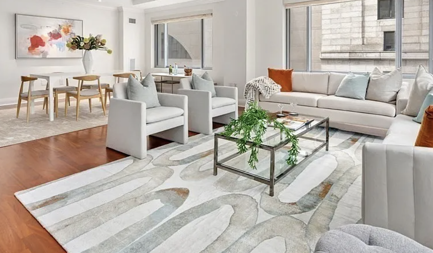 Modern living room with white sofas and armchairs, glass coffee table with green plants, area rug with light abstract pattern, large windows, and a dining table with chairs in the background.