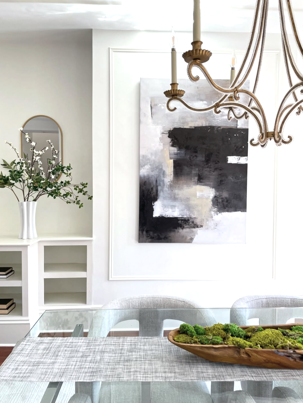 An elegant dining room with glass table, upholstered chairs and neutral large artwork. There is a bronze chandelier in the foreground.
