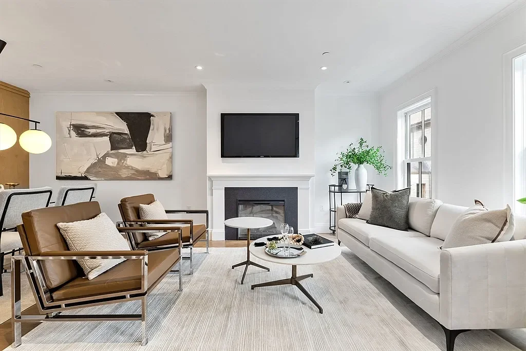 Modern living room with white walls, large windows, beige and white sofas, brown armchairs, a black fireplace, a mounted flat-screen TV, abstract artwork, and decorative plants.