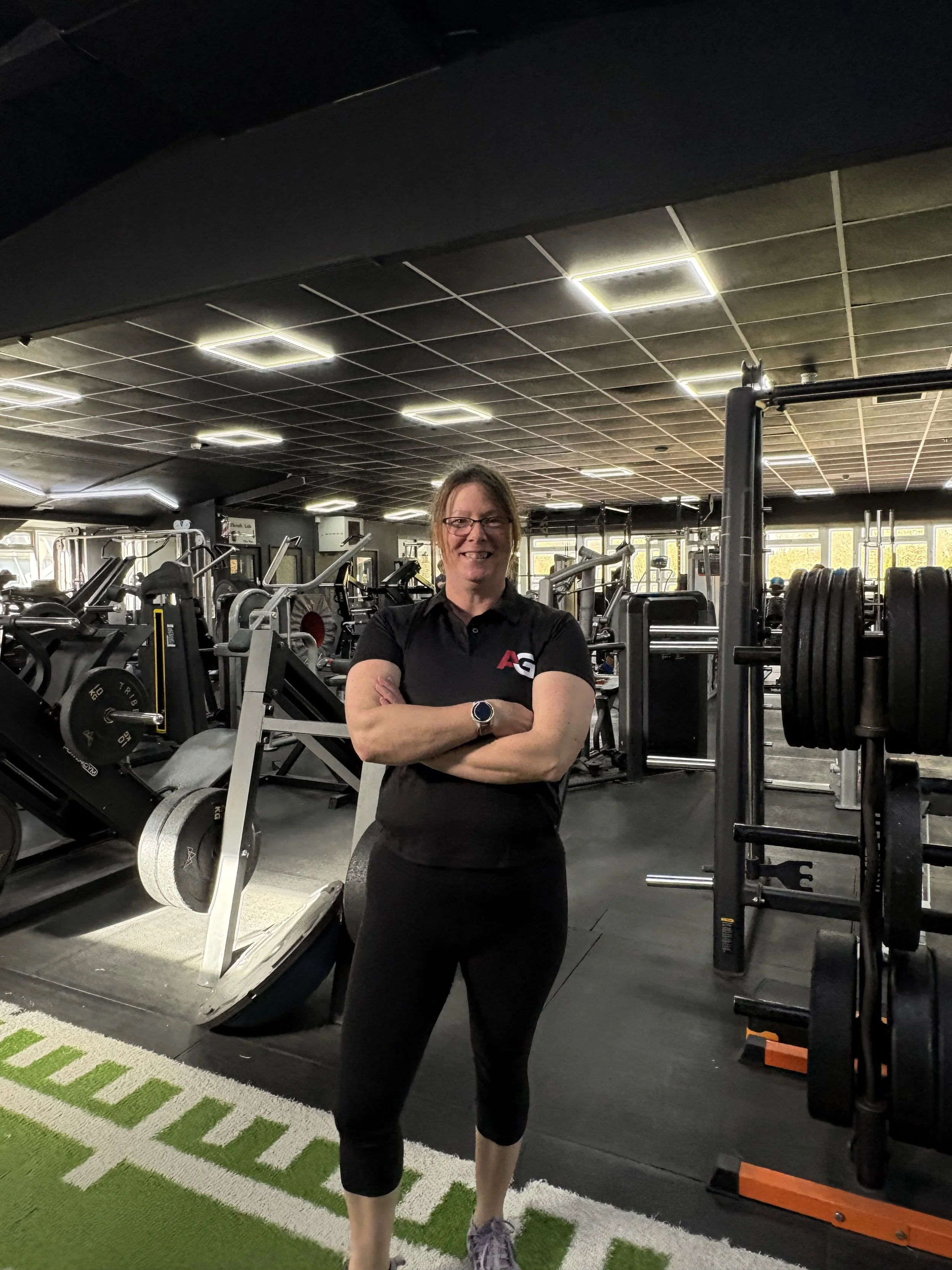 A woman standing with arms crossed inside a gym with various workout equipment and weights.